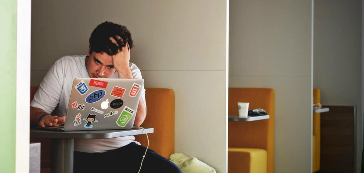 jovem em frente ao seu computador, decorado com muitos autocolantes divertidos, está com a mão na cabeça e com um ar muito preocupado