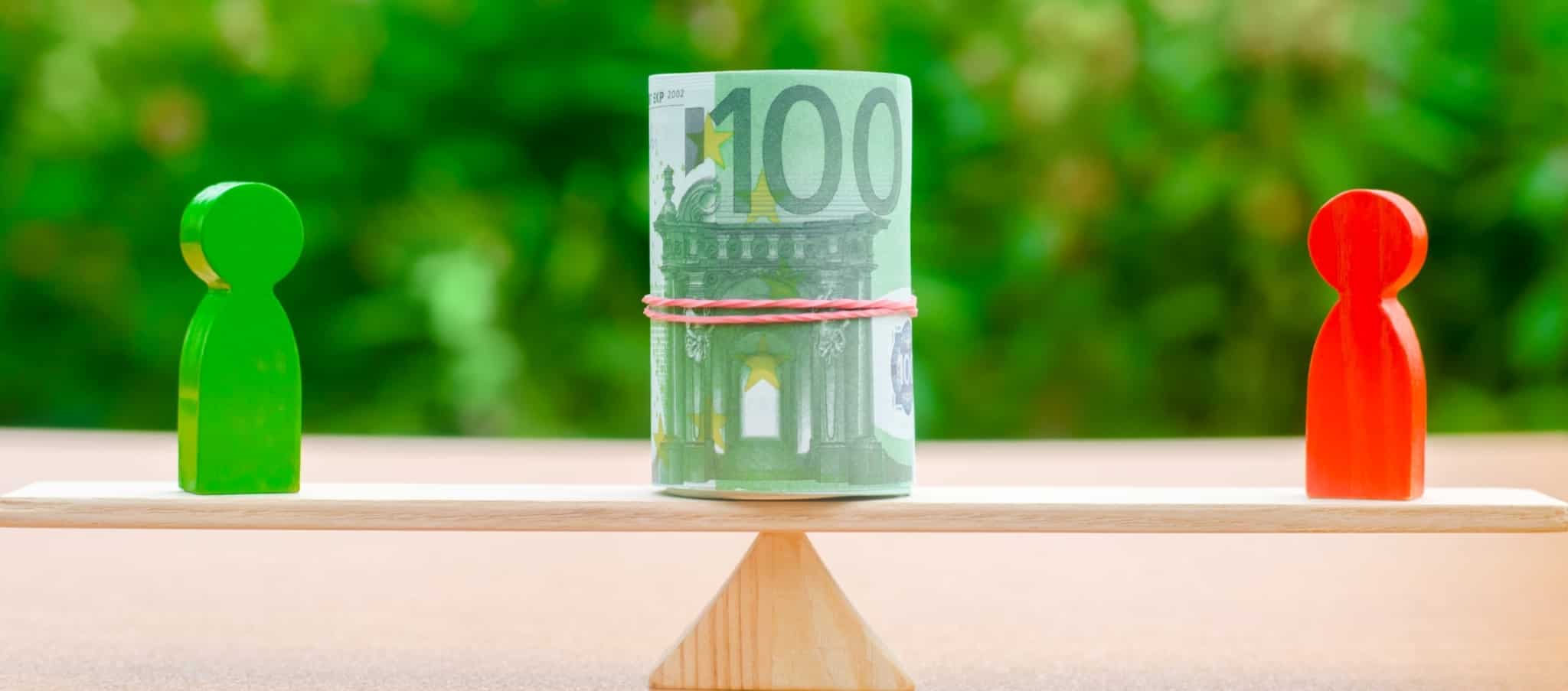 balança de madeira com um rolo de notas de 100 euros e duas figuras em madeira