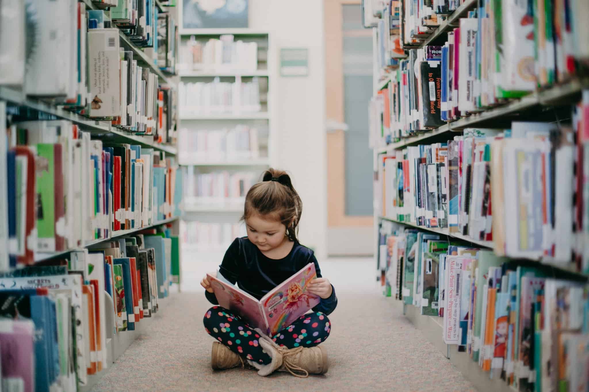 rapariga sentada no chão de uma biblioteca a ler um livro