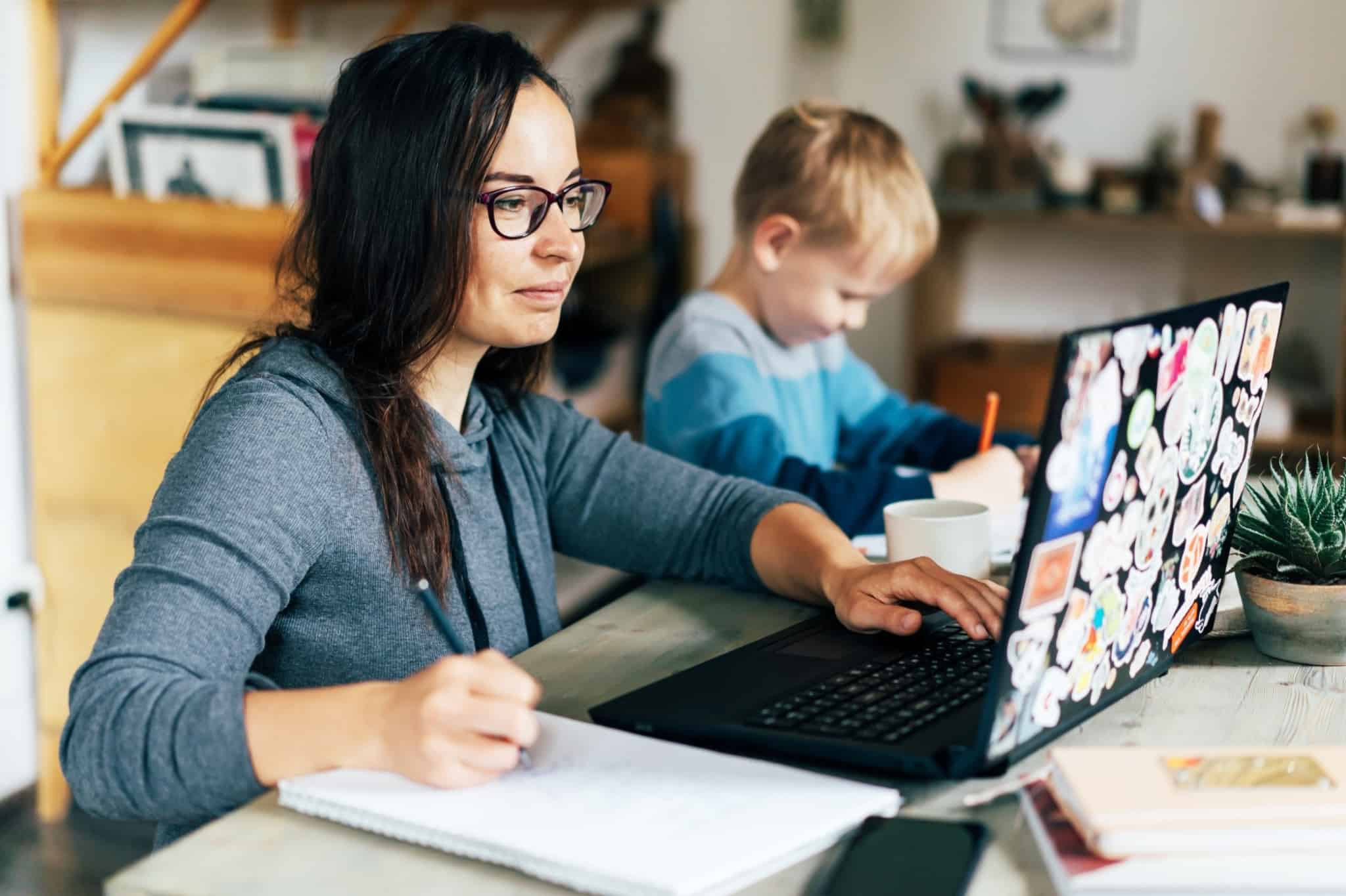 Mulher sentada em frente ao computador com o filho a desenhar ao lado