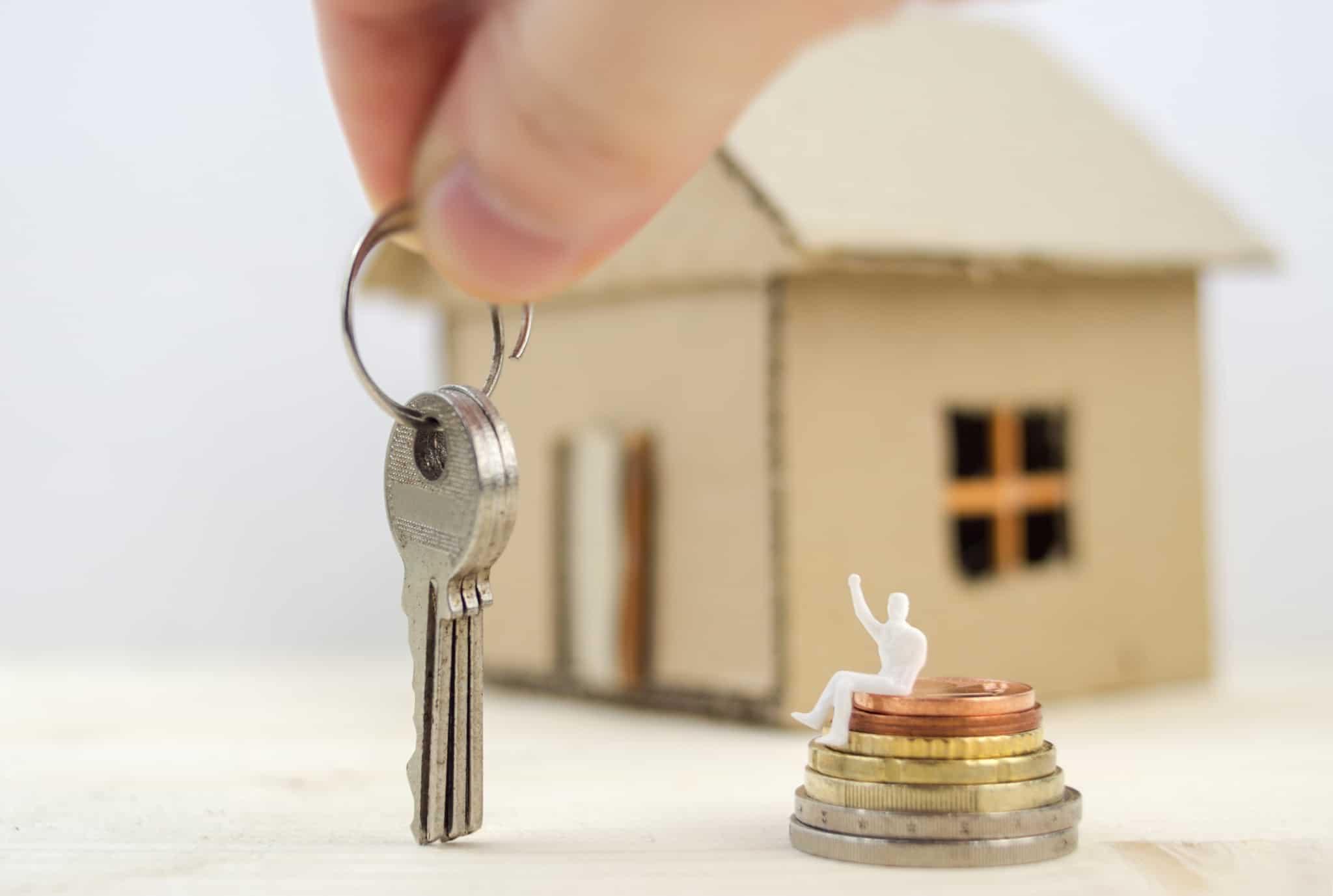 Mão a segurar numa chave de casa ao lado de uma pilha de moedas e uma miniatura de uma casa atrás