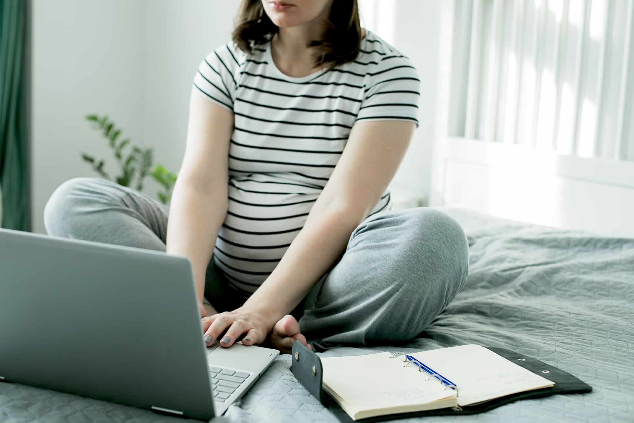 mulher grávida sentada em cima da cama com o computador