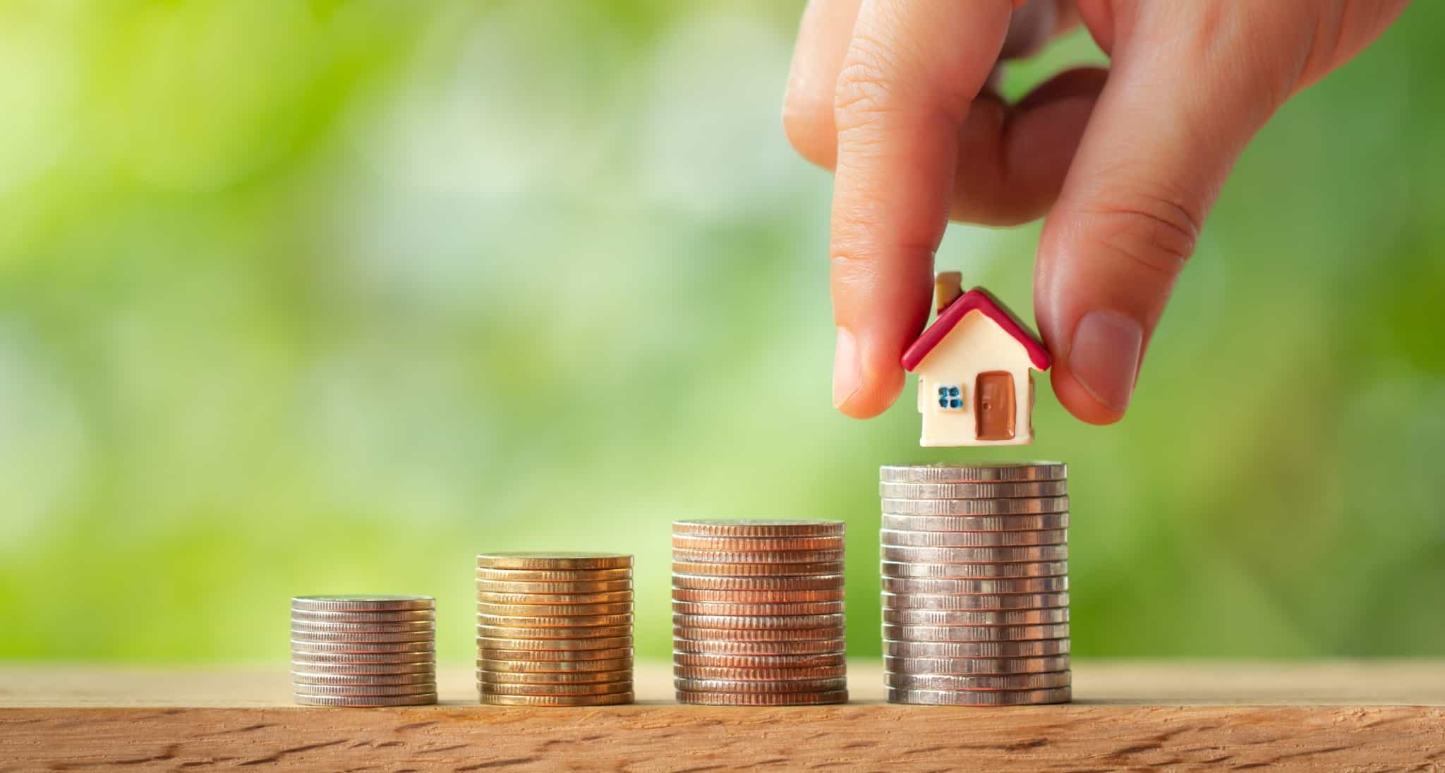 mão a colocar uma casa miniatura numa pilha de moedas