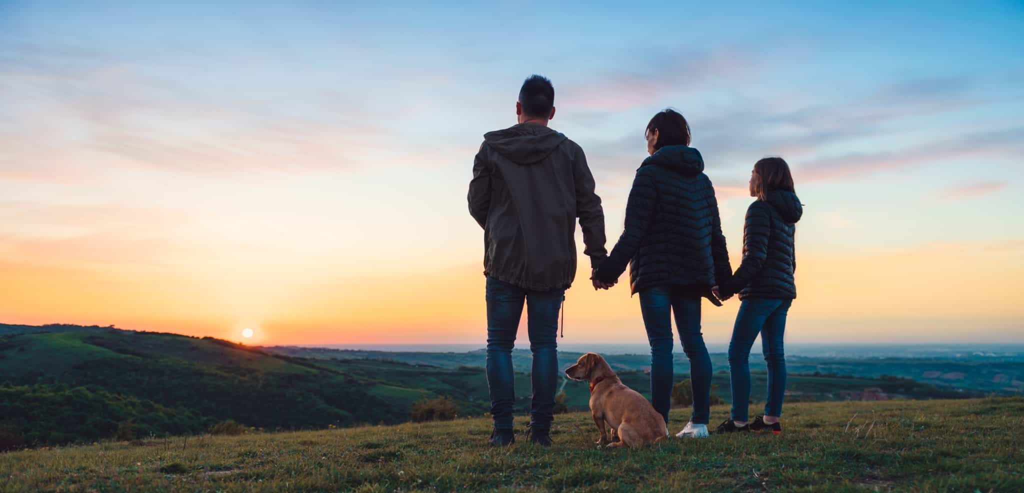 Uma família (pai, mãe e criança) de mãos dadas a ver o pôr do sol, acompanhados pelo cão