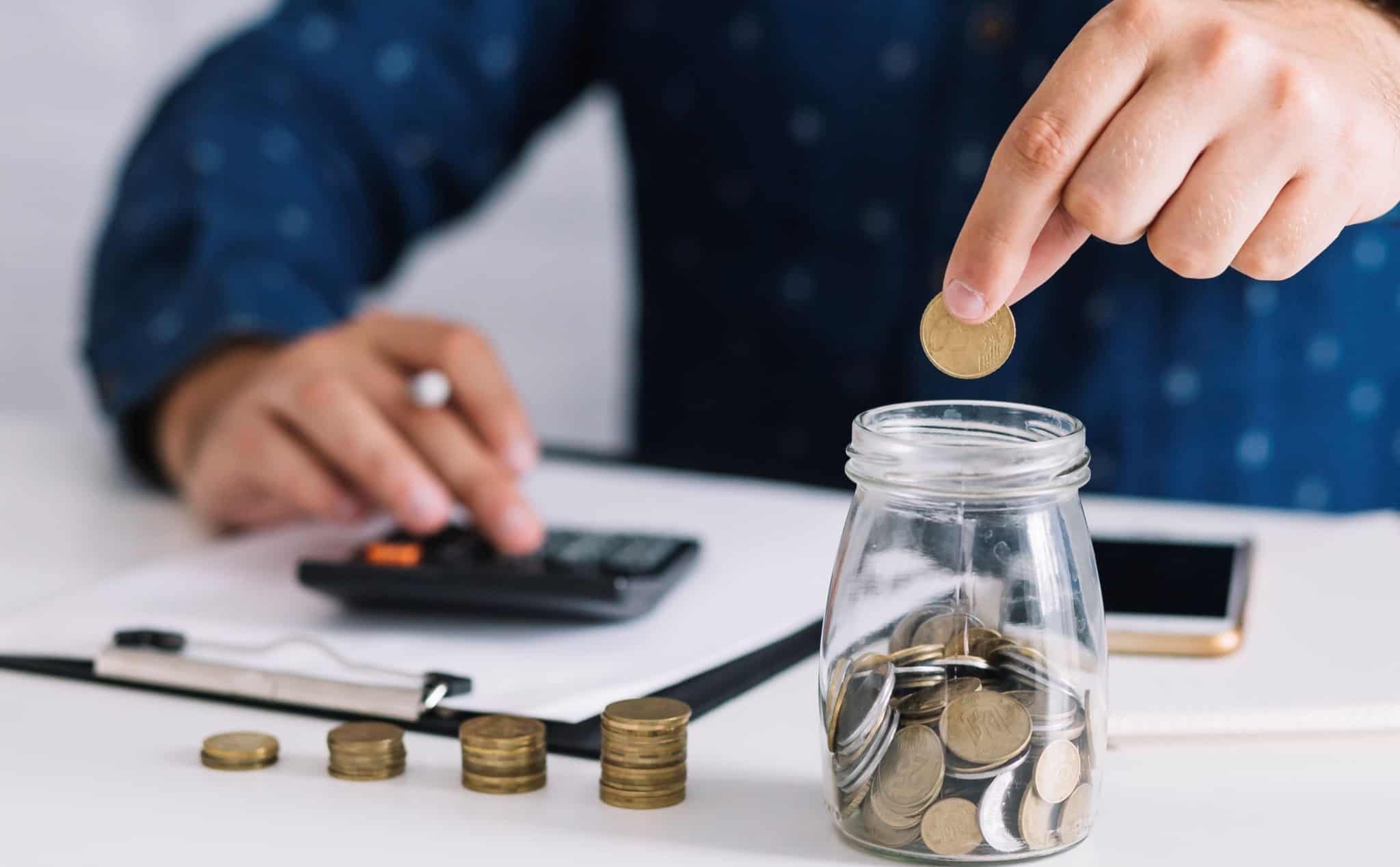 homem está a fazer contas com uma calculadora ao mesmo tempo que coloca umas moedas num mealheiro