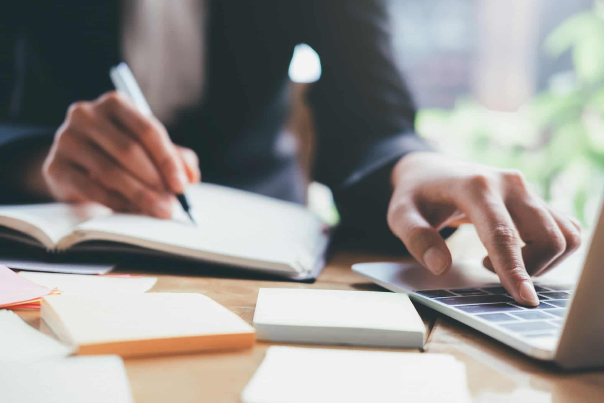 pessoa a escrever num bloco de notas e no computador portatil