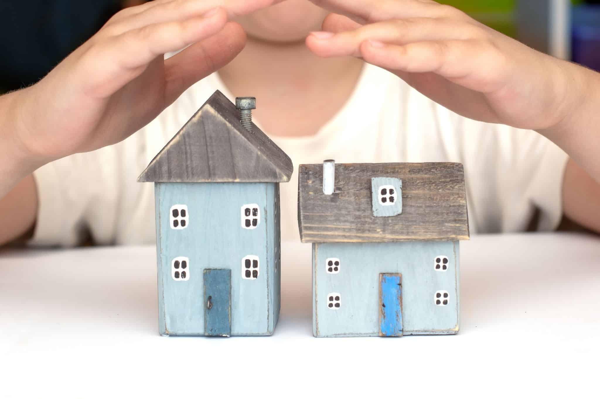 Imagem de duas casas em miniatura com duas mãos a proteger