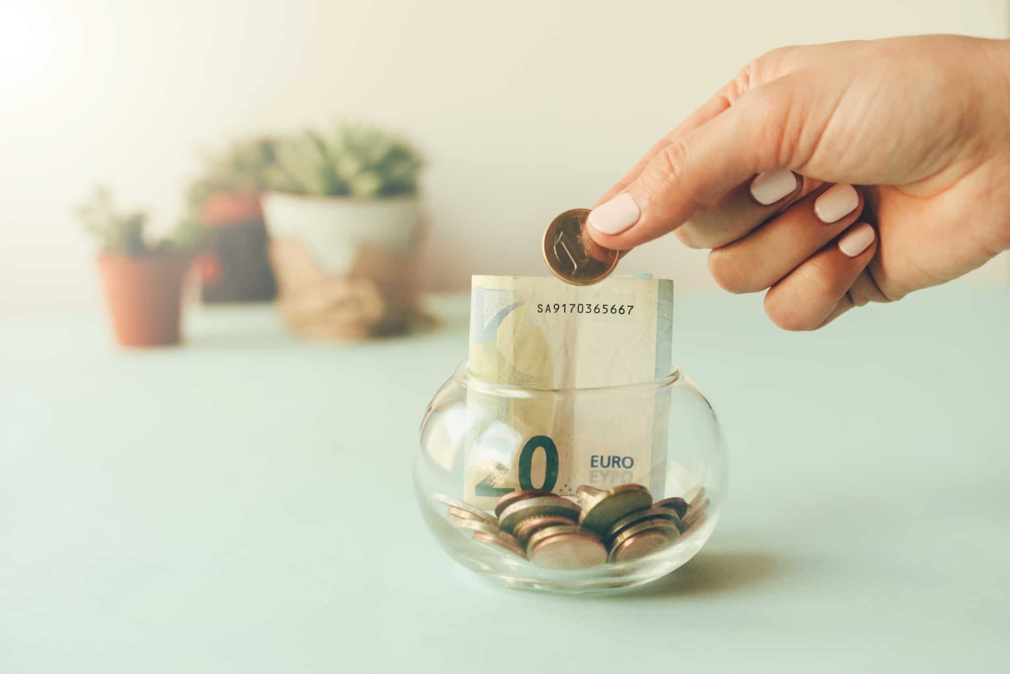 Frasco de vidro com moedas e notas e uma mão a colocar mais uma moeda de 1 euros