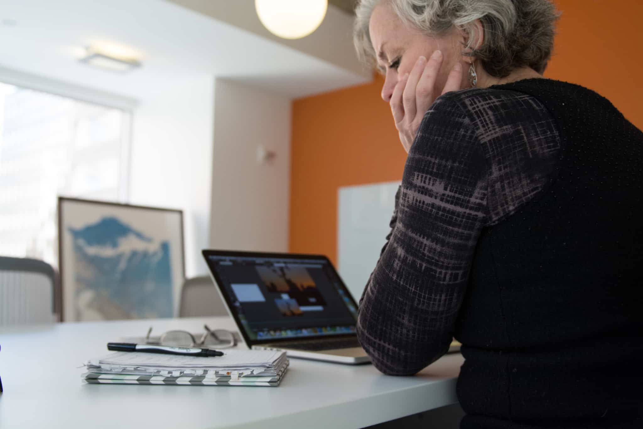 Mulher de cabelo grisalho consulta uns apontamentos num caderno, enquanto trabalha num computador