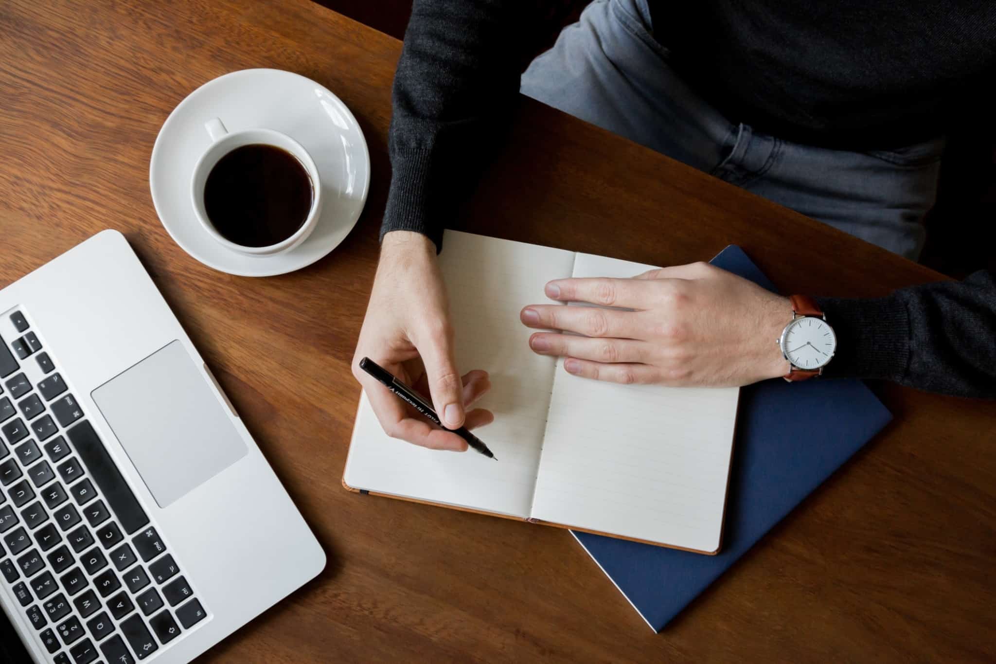 Pessoa a anotar despesas num caderno. À sua frente está um computador e uma chávena de café