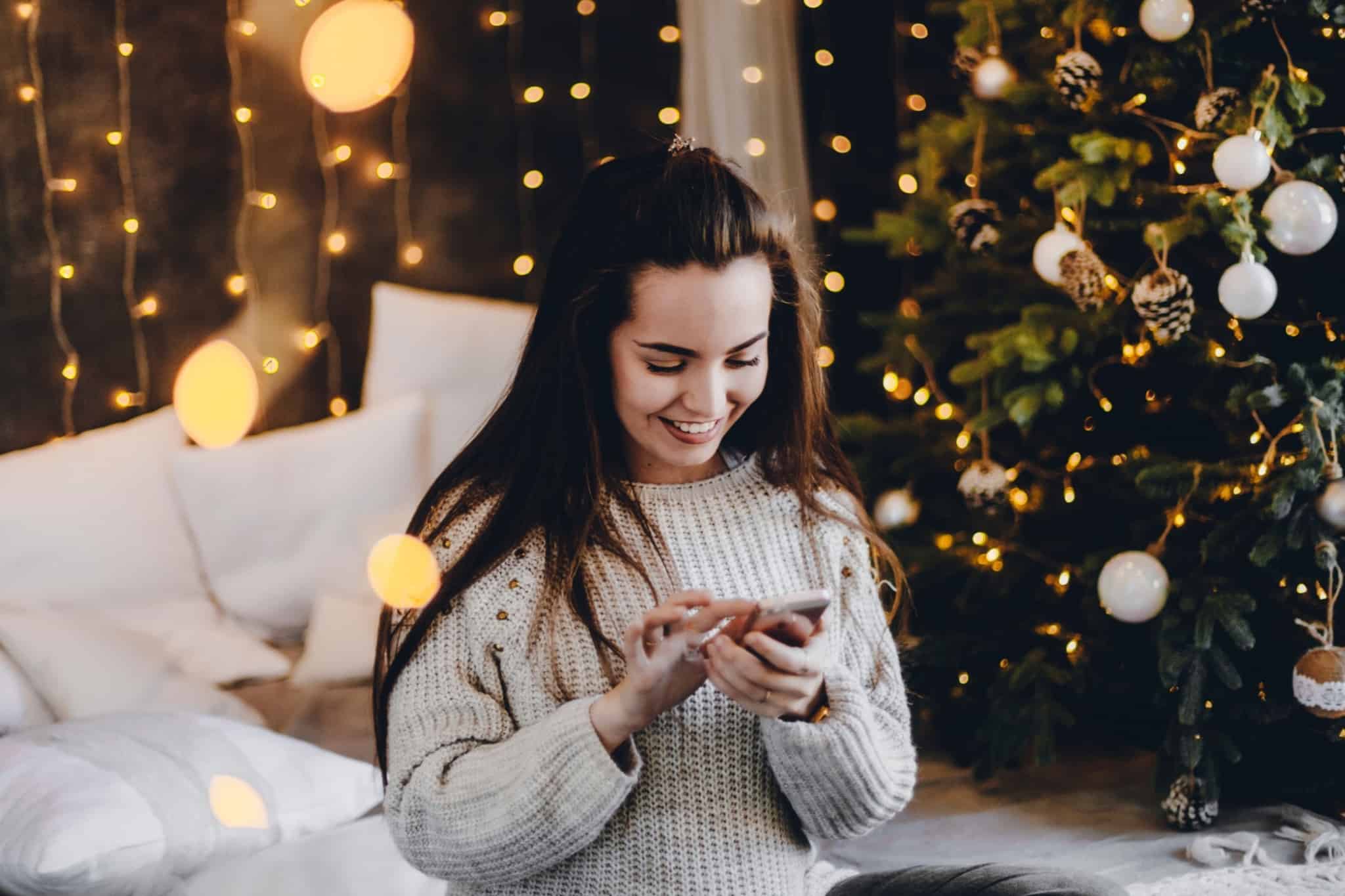 Rapariga a sorrir, com o telemóvel na mão e a árvore de Natal iluminada atrás