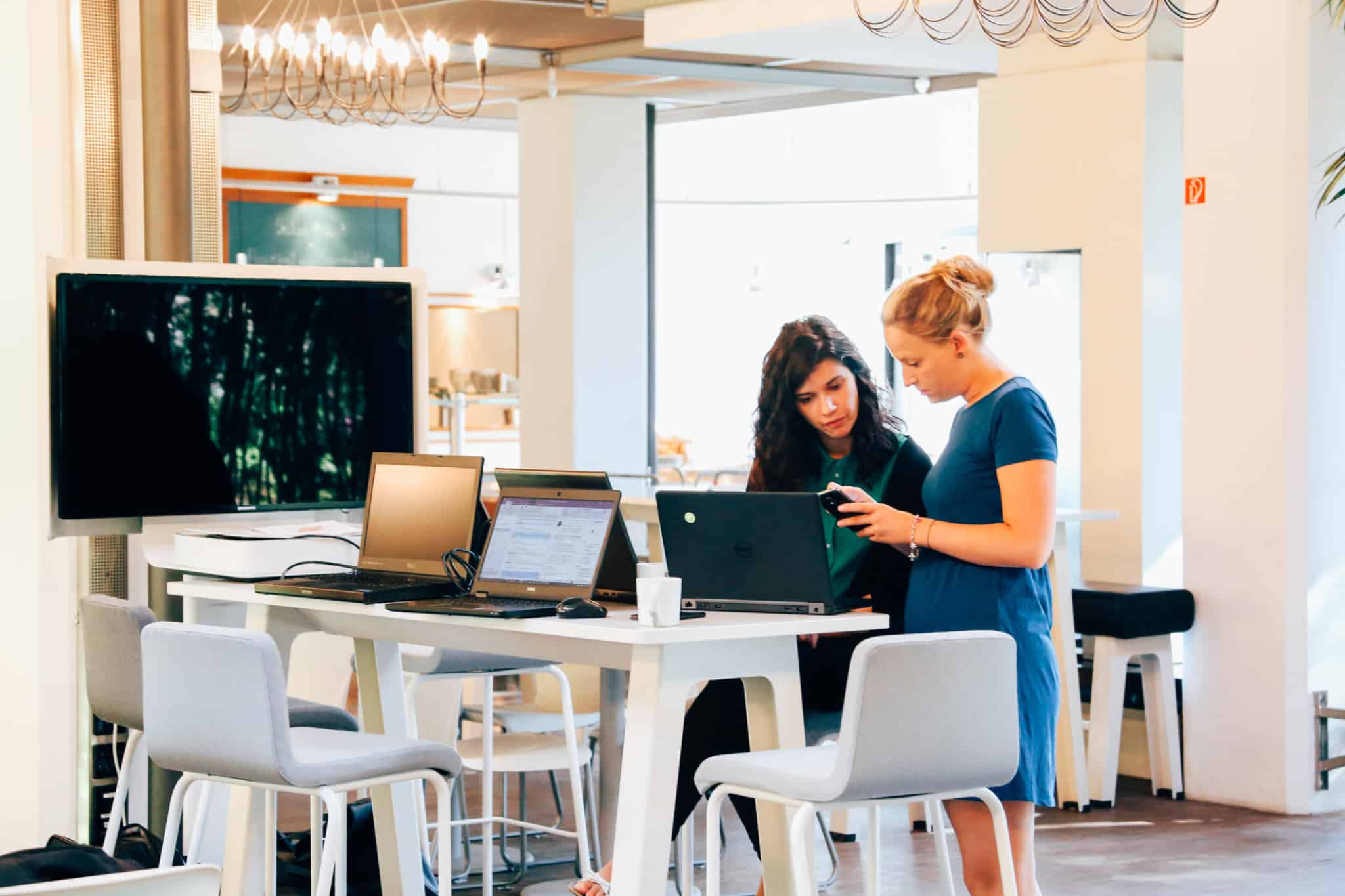 Duas mulheres conversam junto a uma mesa alta em frente a computadores e aos telemóveis