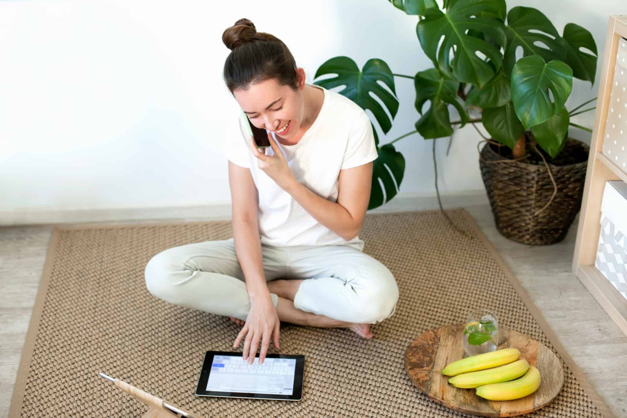 Mulher sentada no chão de casa a falar ao telemóvel ao mesmo tempo que consulta um tablet. À sua frente tem um prato com bananas