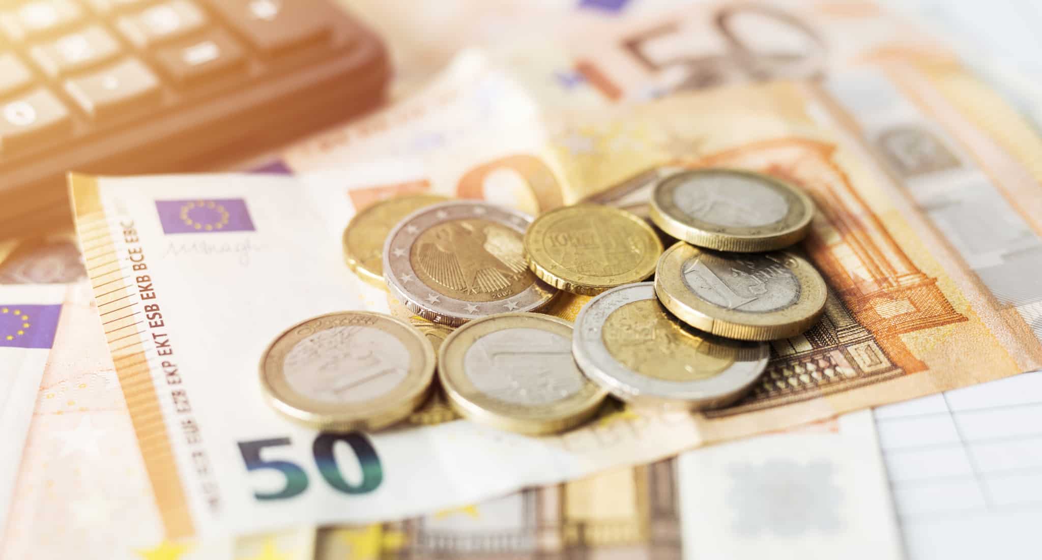 várias moedas de euro em cima de uma nota de 50 euros. Ao lado uma calculadora