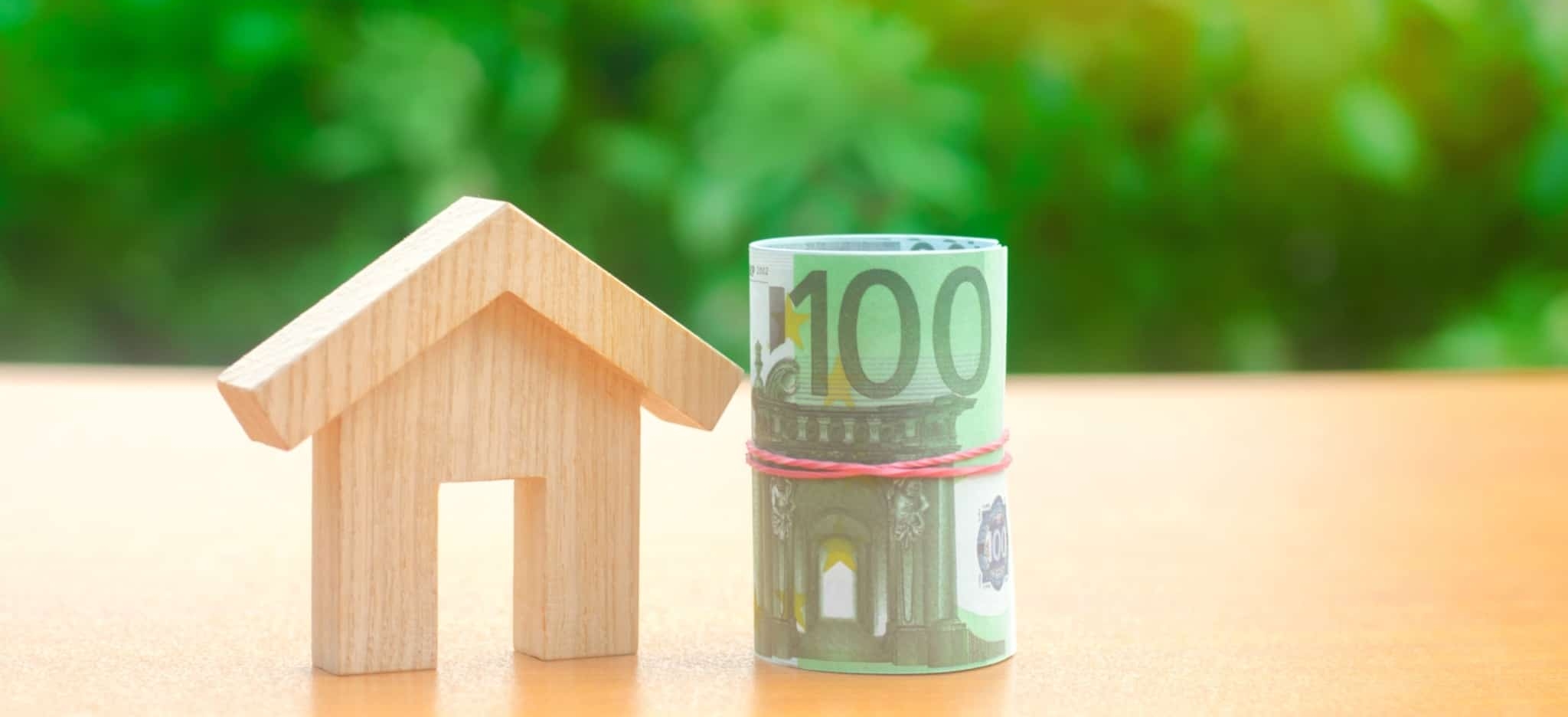 Miniatura de uma casa em madeira ao lado de um rolo de notas de 100 euros