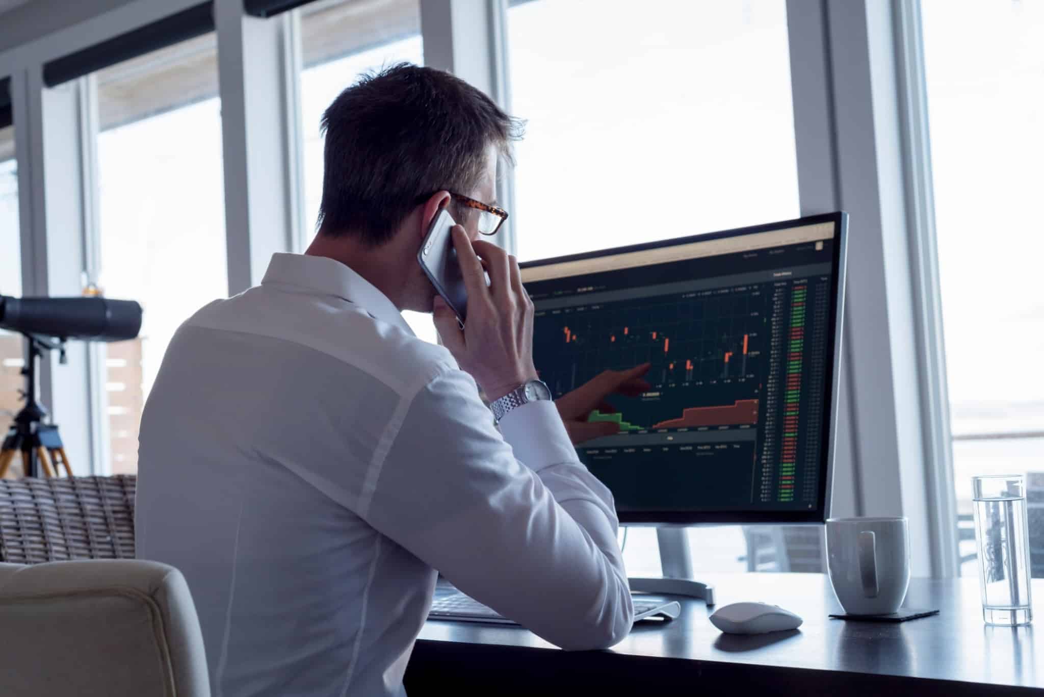 Investidor fala ao telemóvel ao mesmo tempo que analisa um gráfico no computador