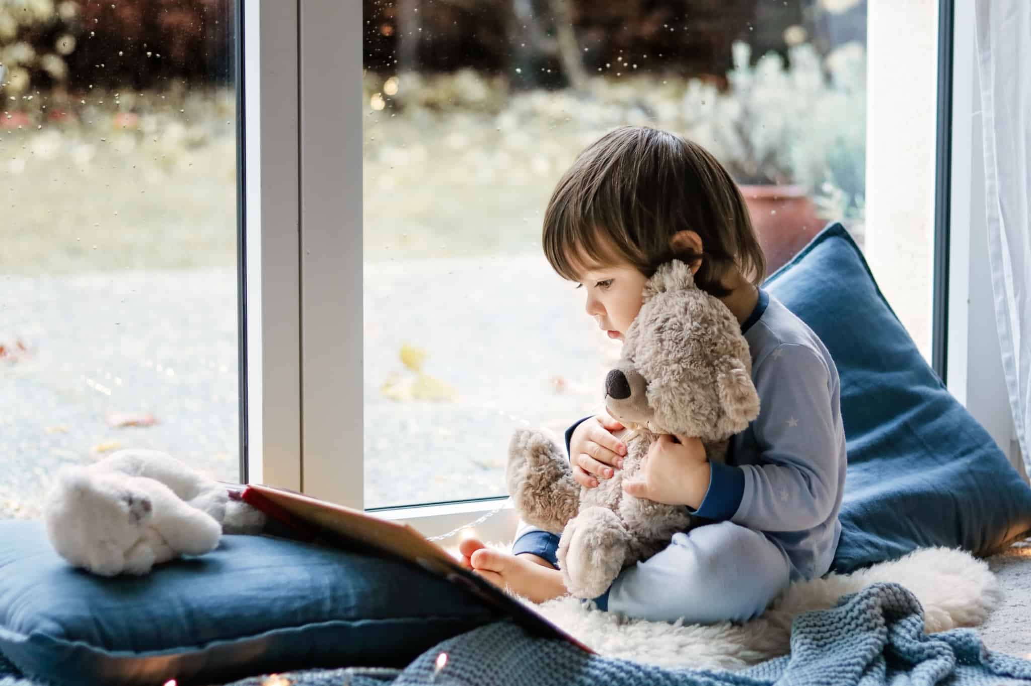 Criança está junto a uma janela a ver um livro enquanto agarra um urso de peluche