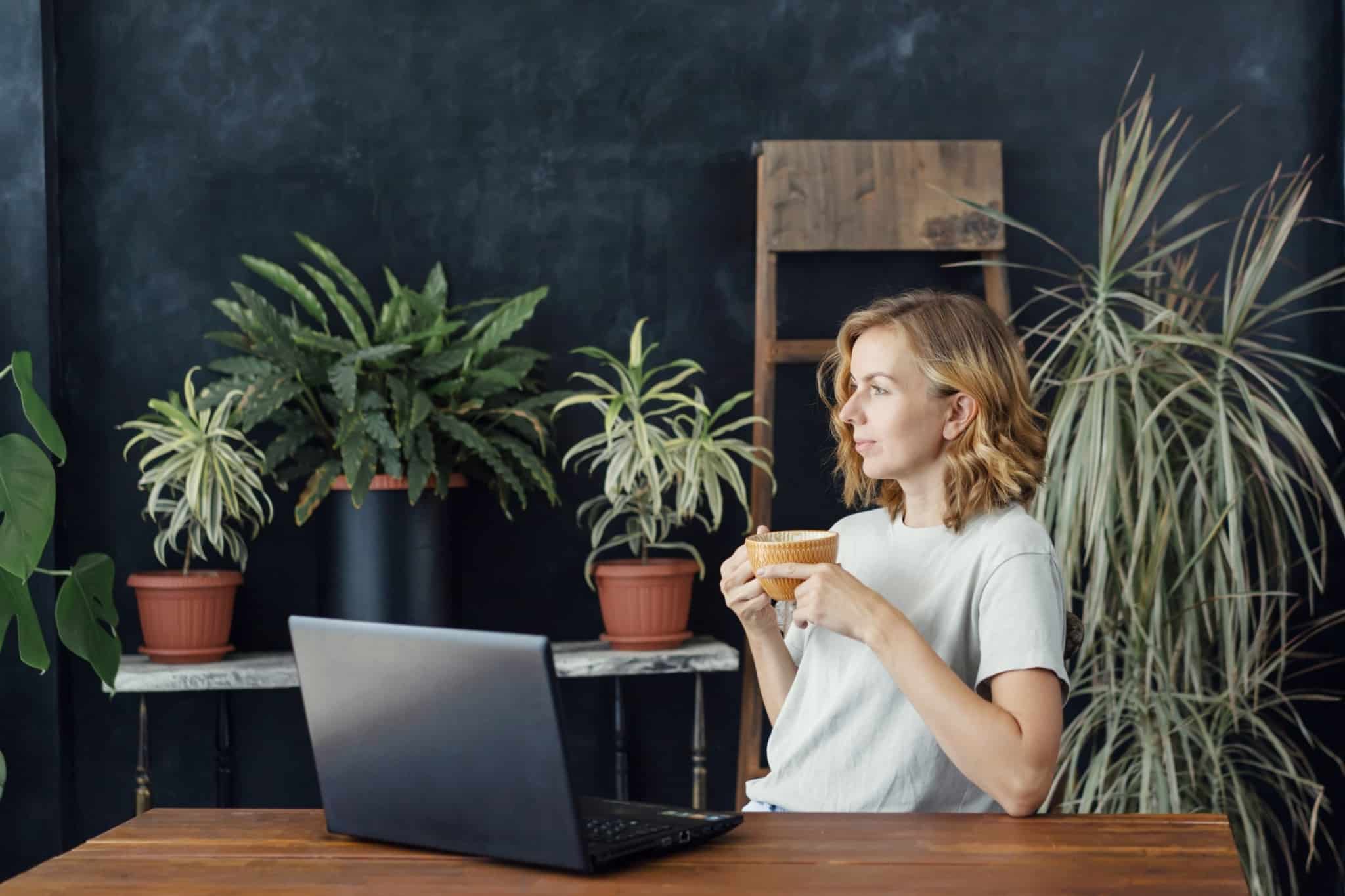 Mulher sentada a beber café e com o computador à sua frente. Atrás várias plantas