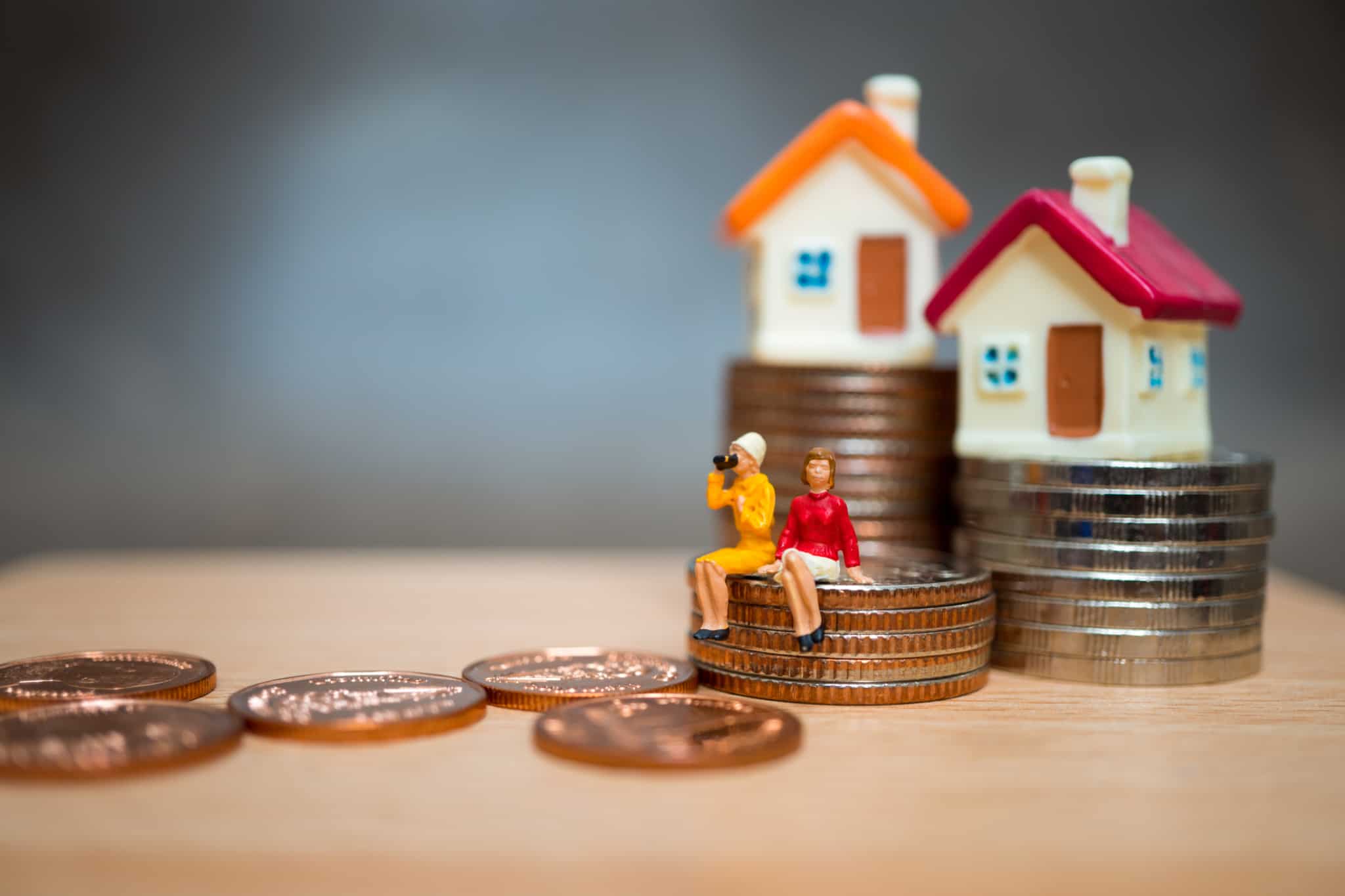 bonecos de miniatura sentados em cima de uma pilha de moedas. Ao fundo umas casa miniaturas em cima de duas pilhas de moedas