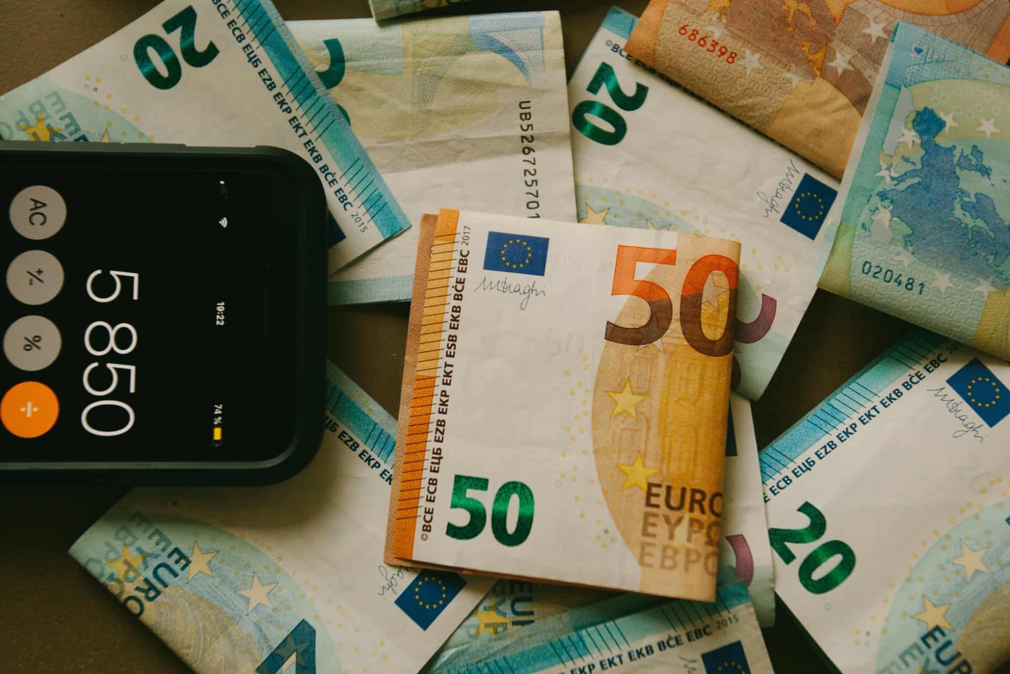 várias notas de euro espalhadas em cima de uma mesa e ao lado um telemóvel com a máquina de calcular ativada