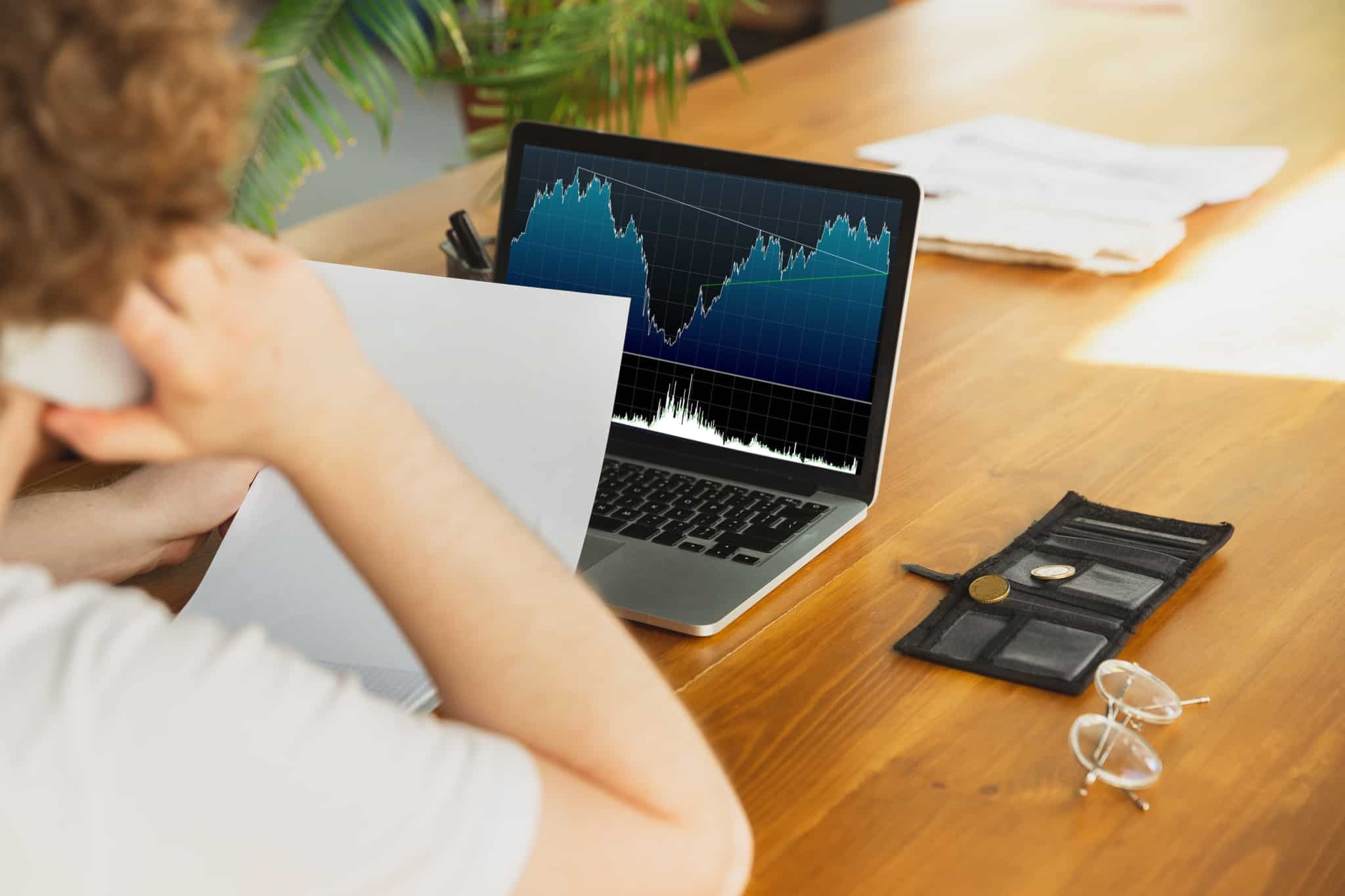 Homem ao telefone enquanto analisa o desempenho dos seus investimentos, através de uma folha e de um gráfico no computador