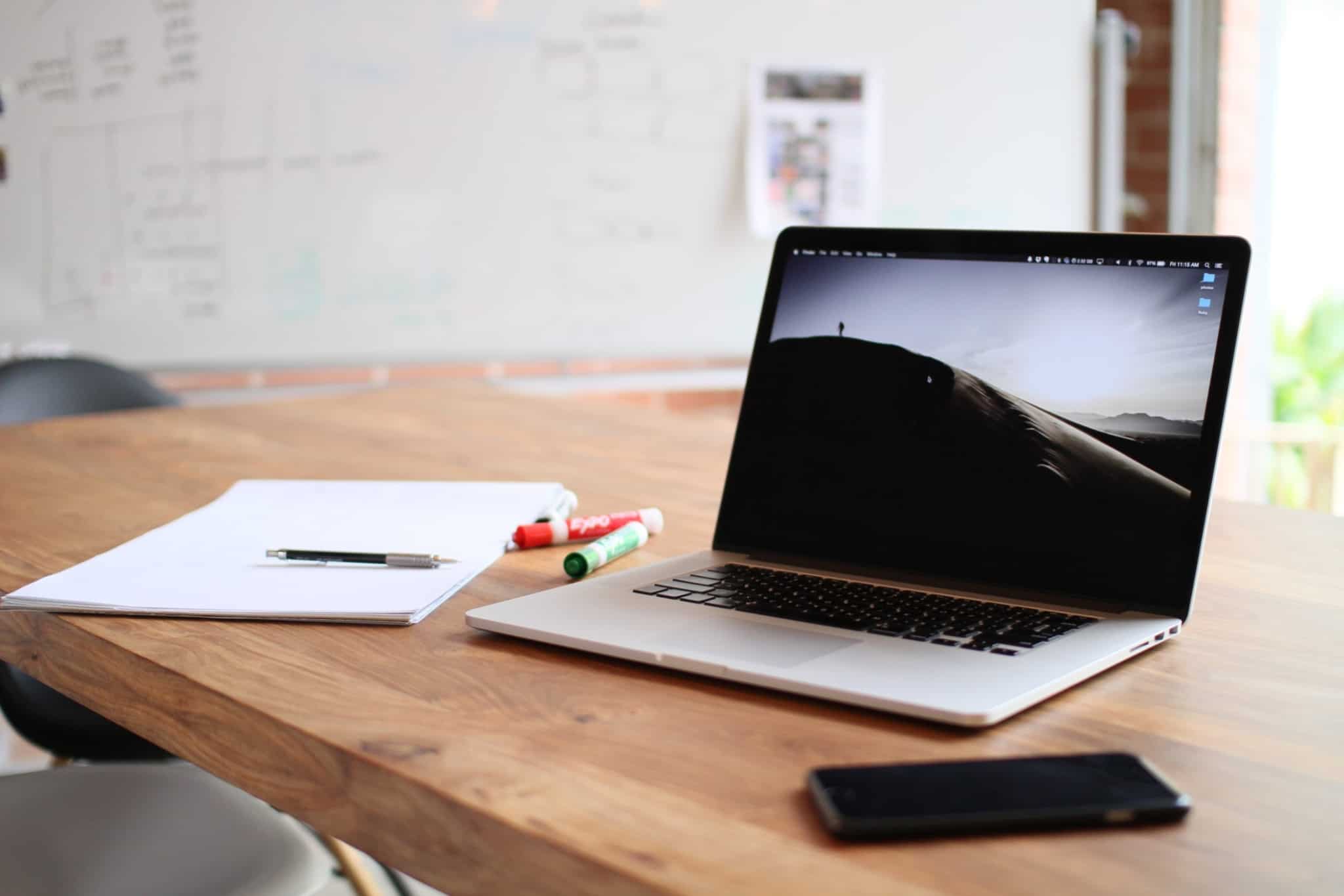 Computador em cima de uma mesa, ao pé de um bloco de notas de um telemóvel e de uns marcadores