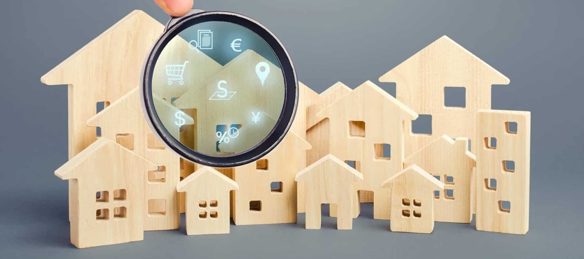 Figuras de casas e prédios em madeira e uma lupa à frente através da qual se veem simbolos de dinheiro, localização e compras