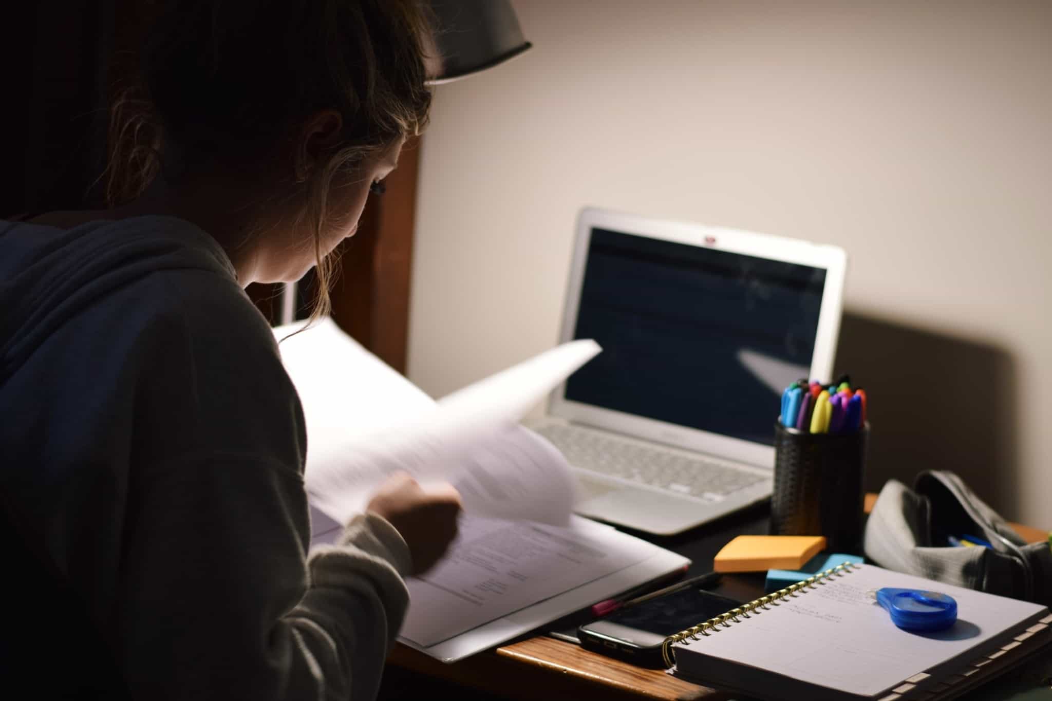 Jovem está a estudar, recorrendo a um livro e com o computador na sua frente