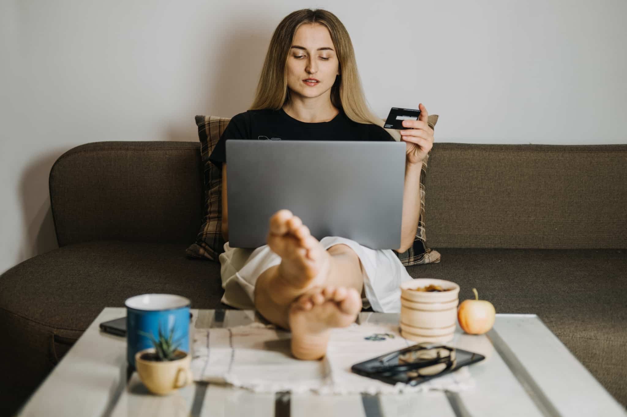 jovem mulher, sentada no sofá com os pés em cima da mesa de centro, tem o computador no colo enquanto o usa para compras online