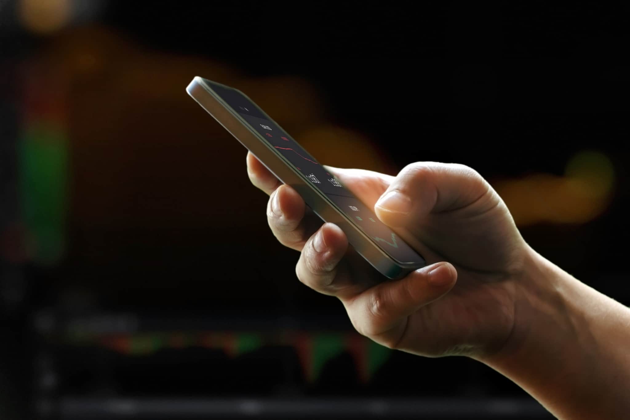 Mão de homem a segurar o telemóvel, através do qual analisa os seus investimentos realizados através de plataformas peer-to-peer