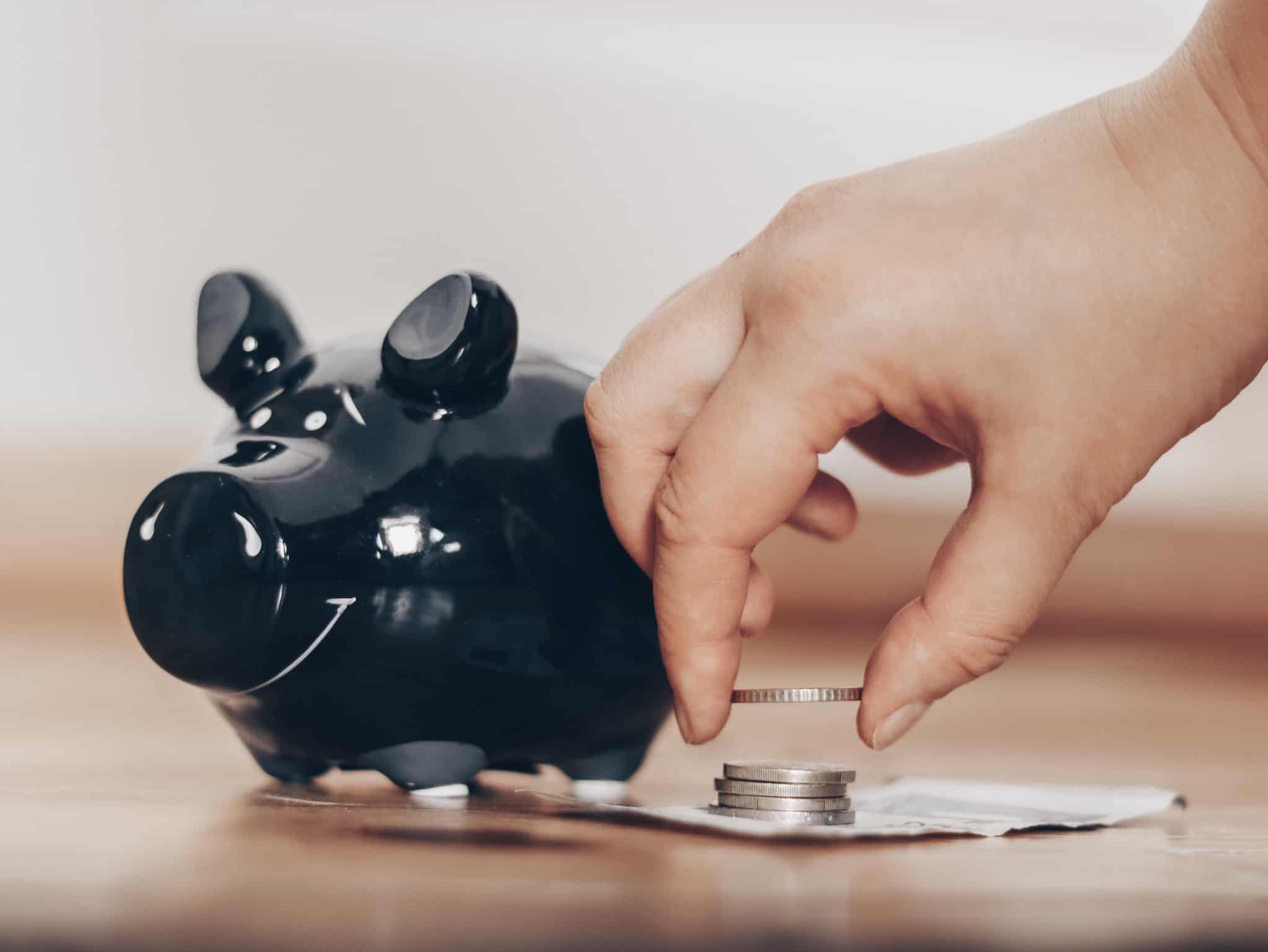 mealheiro em forma de porco. ao seu lado uma mão a pegar em moedas. Usar o subsídio de férias para poupar
