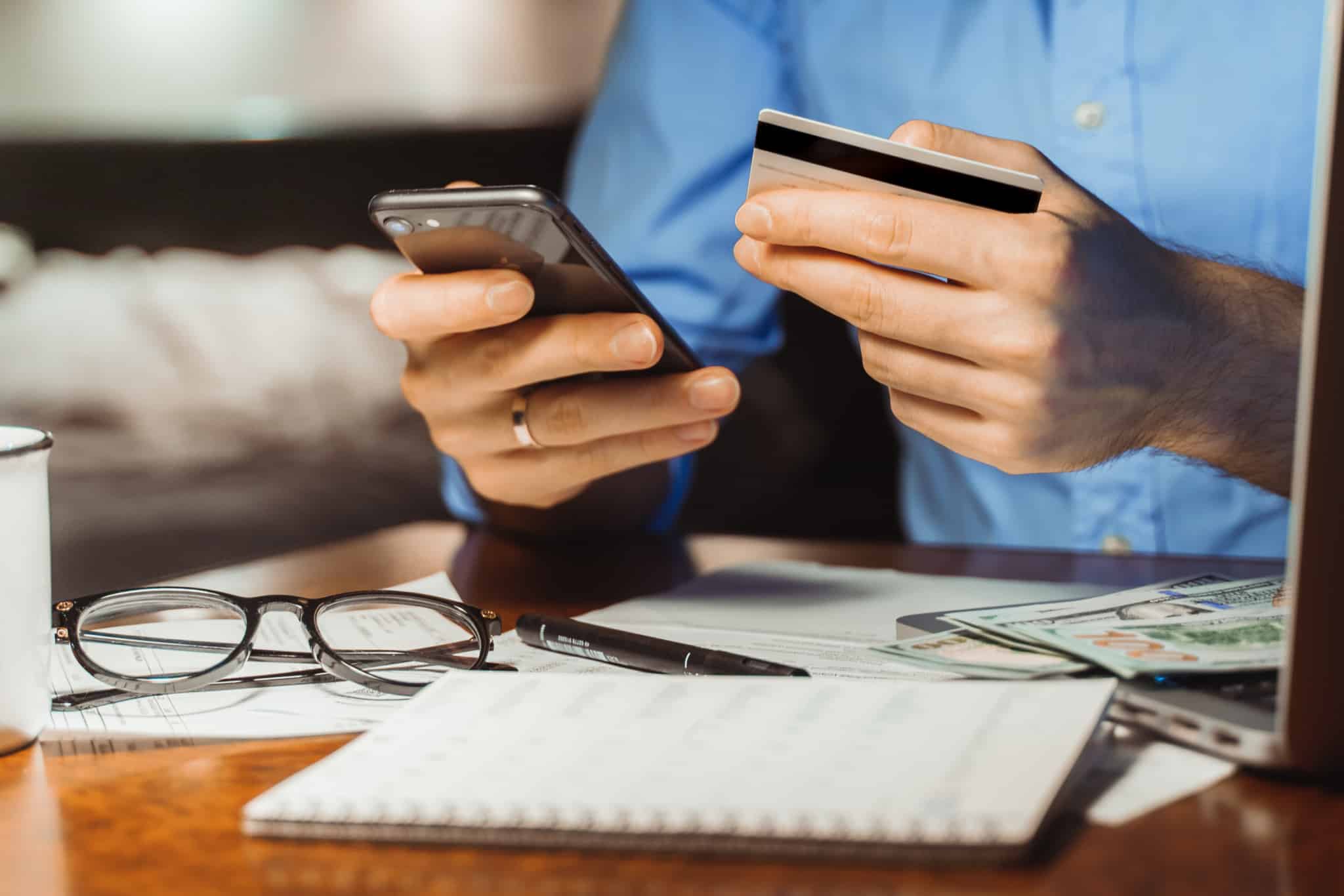 Homem consulta a sua conta bancária no telemóvel. Na outra mão tem um cartão de crédito