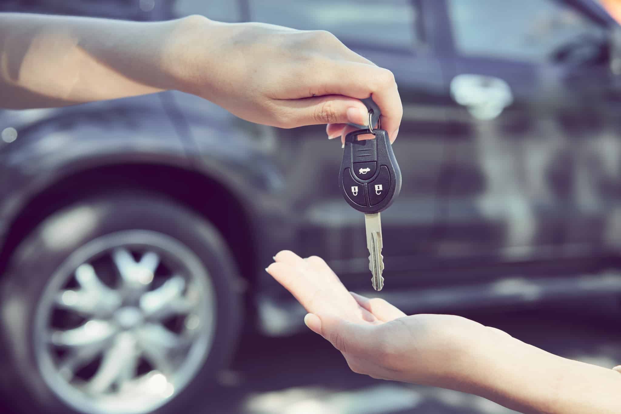 grande plano de duas mãos com um carro como pano de fundo, mostrando a entrega de chaves e simbolizando a compra do veículo.