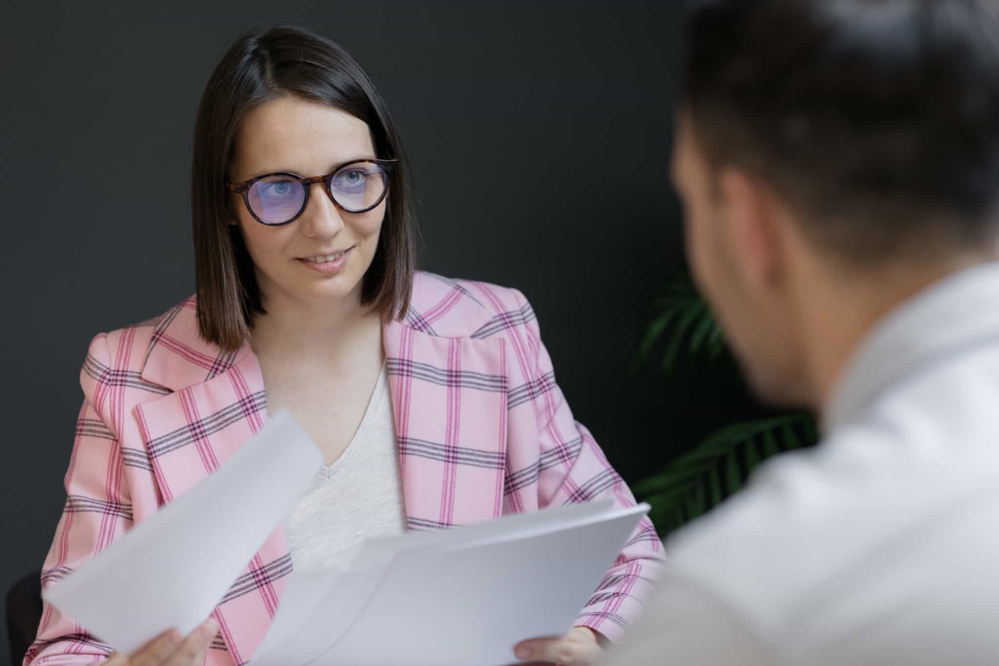 Jovem mulher entrevista, num processo de recrutamento,, um jovem rapaz. Ela analisa o currículo para decidir se ele passa às próximas fases de uma entrevista de emprego.