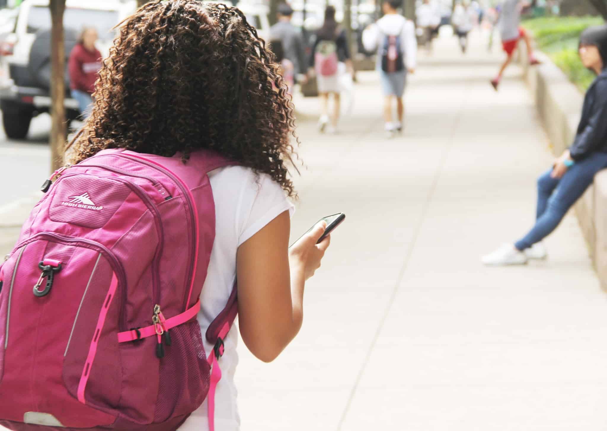 jovem menina leva mochila, cor de rosa, às costas, e encaminha-se, por entre outros jovens, para a sua escola.