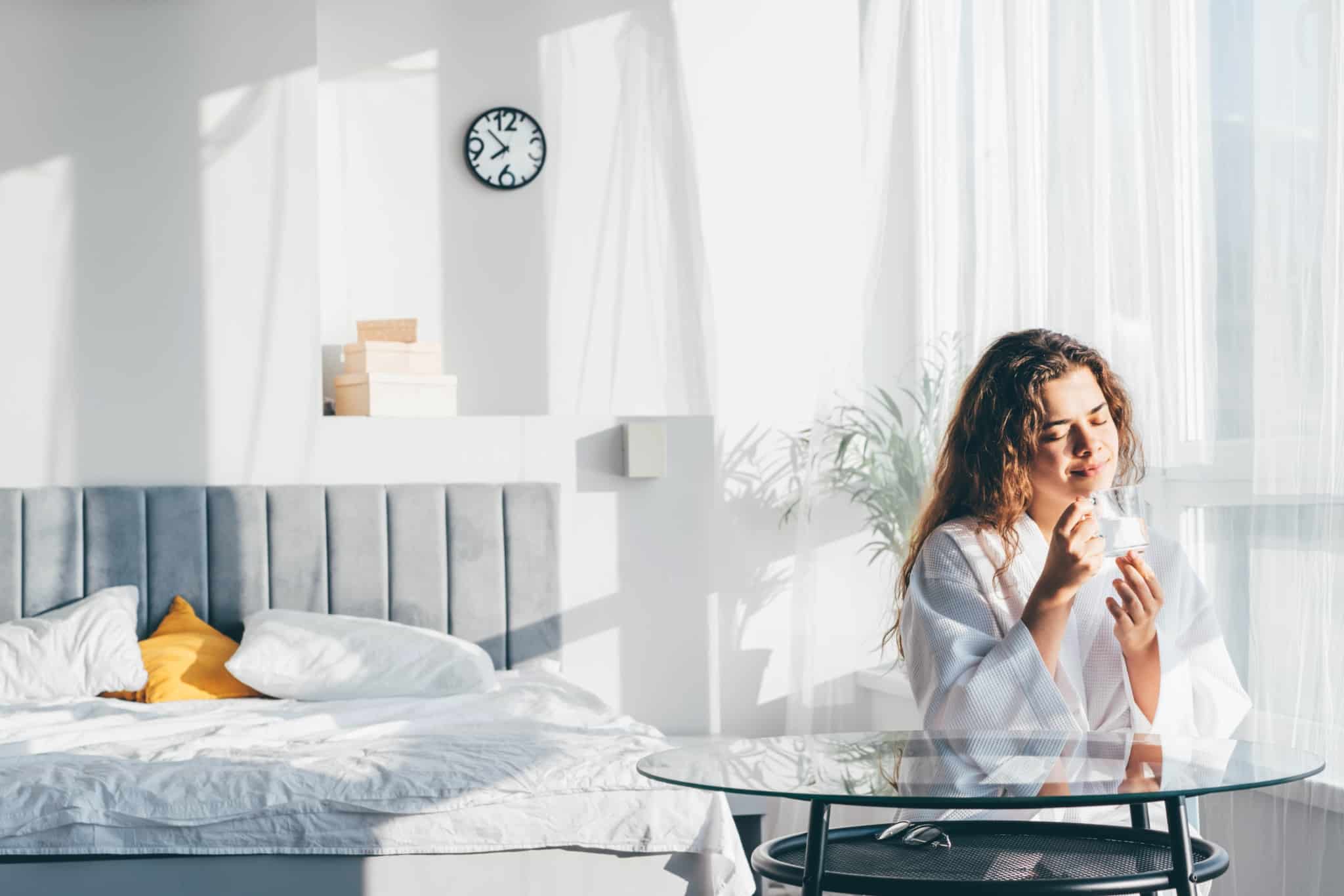jovem mulher, num quarto muito iluminado pela luz natural do romper do dia, toma o seu pequeno almoço em casa