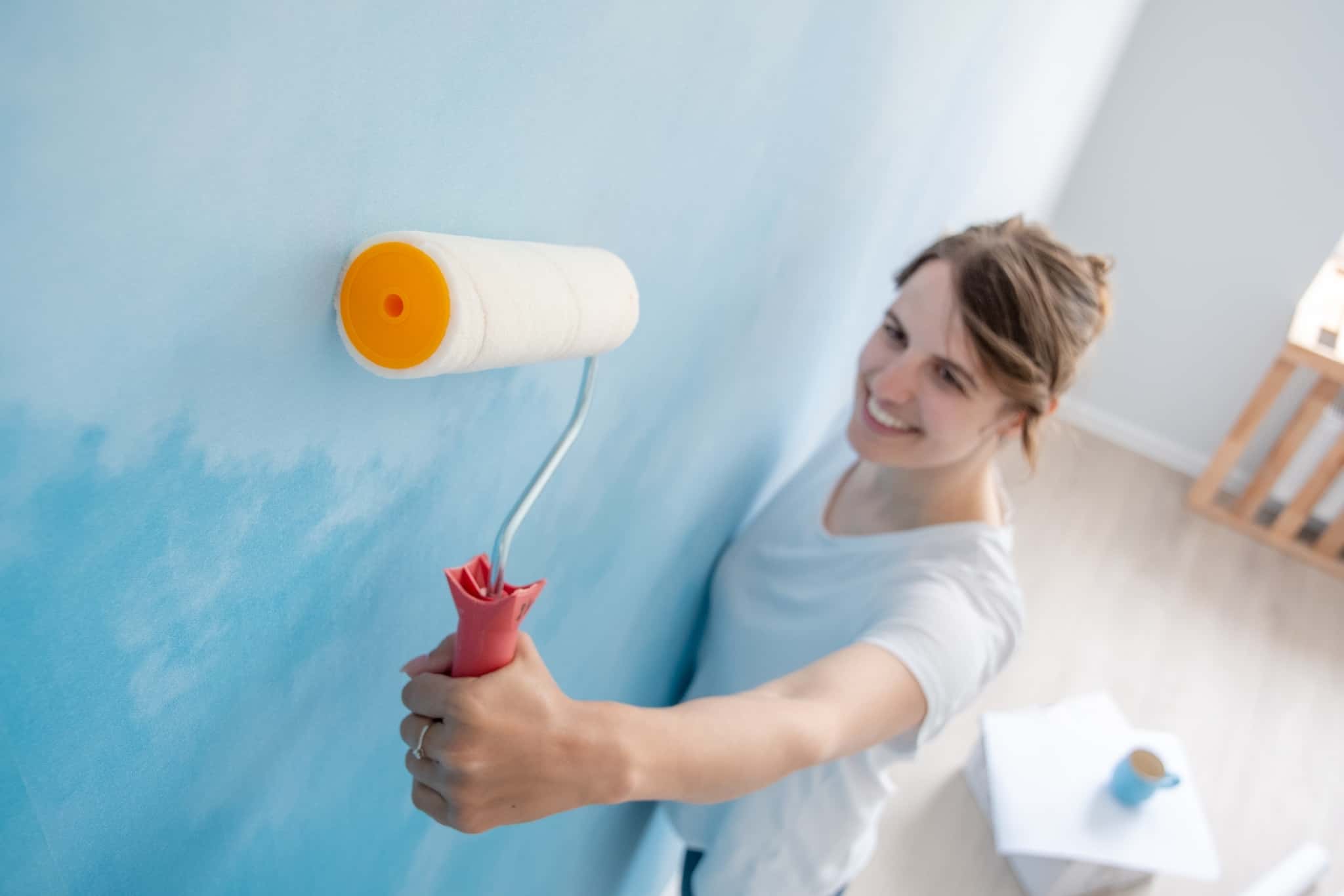 jovem mulher, de rolo de pintar paredes em punho, está em cima de um escadote, enquanto está a remodelar a casa e, assim, ainda consegue poupar.