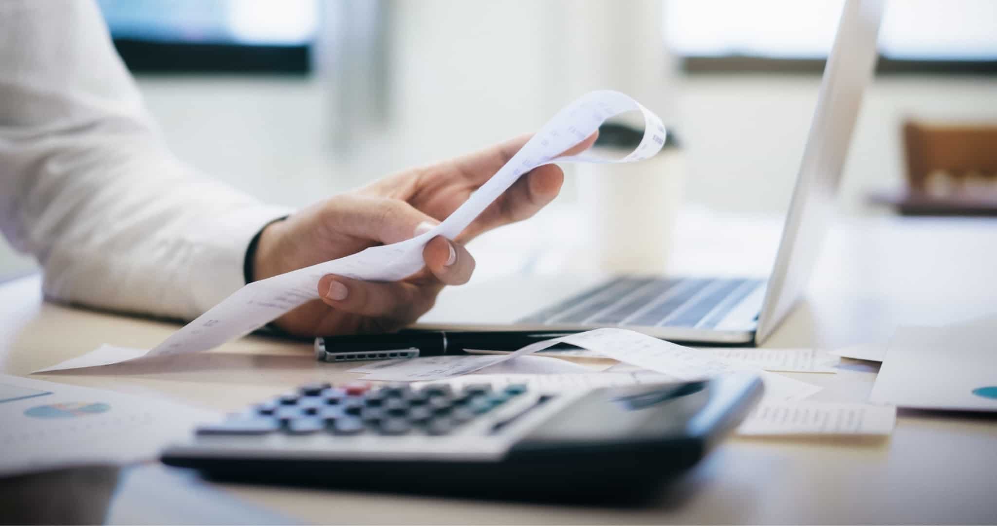 Mão de senhor a segurar um talão com uma calculadora e um portátil à frente a reequilibrar contas