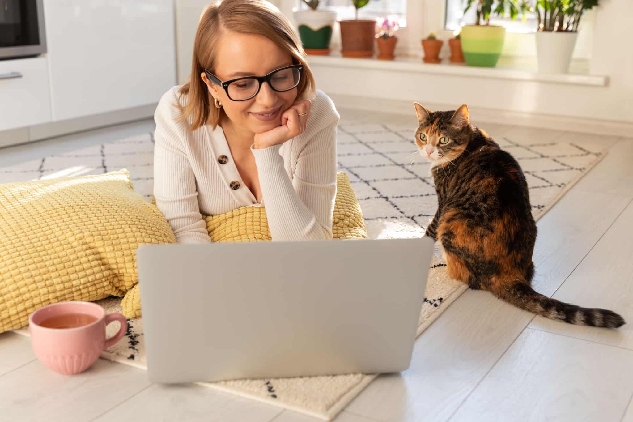 jovem mulher, muito sorridente, trabalha ao computador no chão da sua sala de estar, acompanhada do seu gato, usufruindo dos benefícios flexíveis no trabalho.
