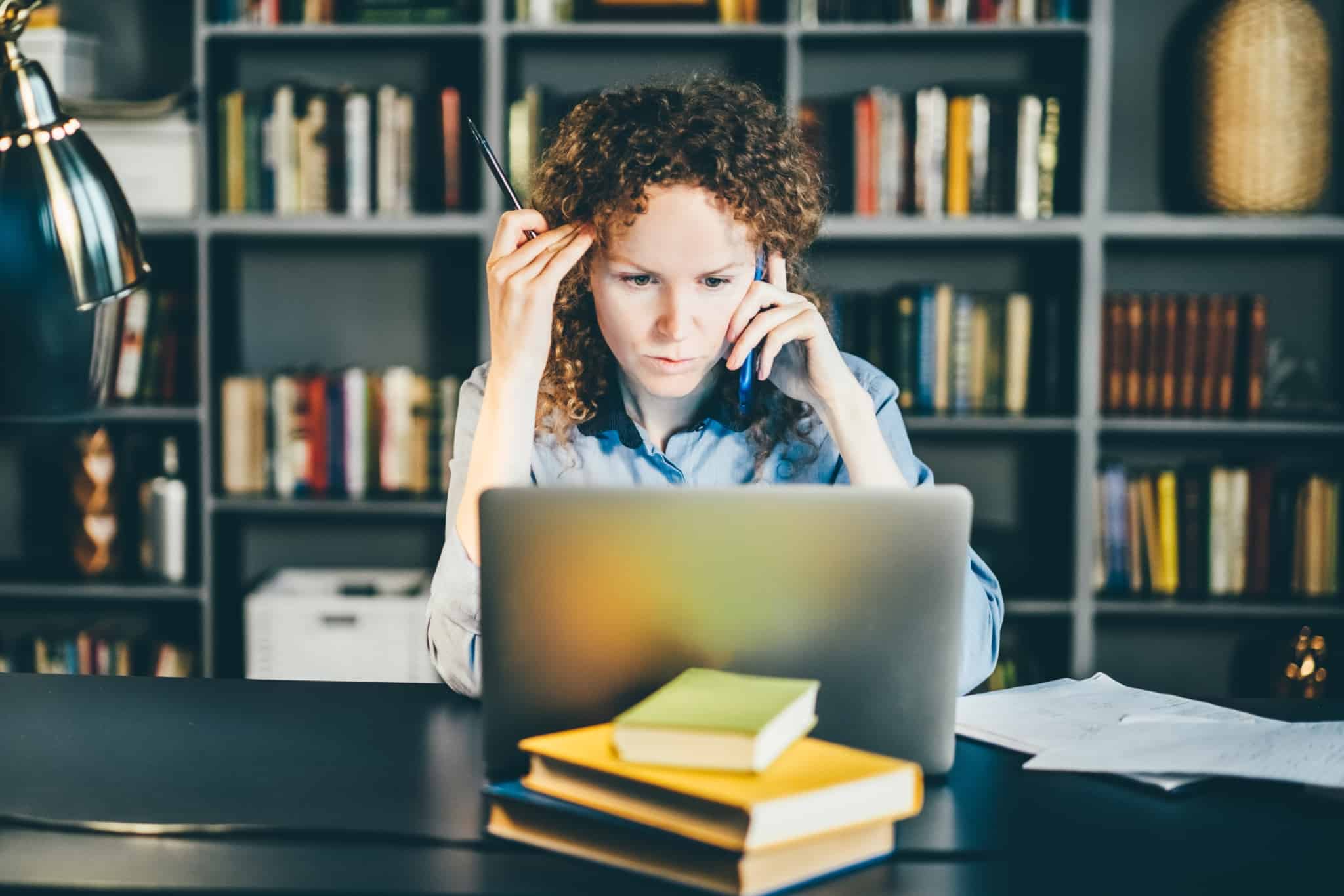 jovem mulher, ao computador e rodeada de papelada, está com um ar muito preocupada, ao analisar os seus investimentos financeiros