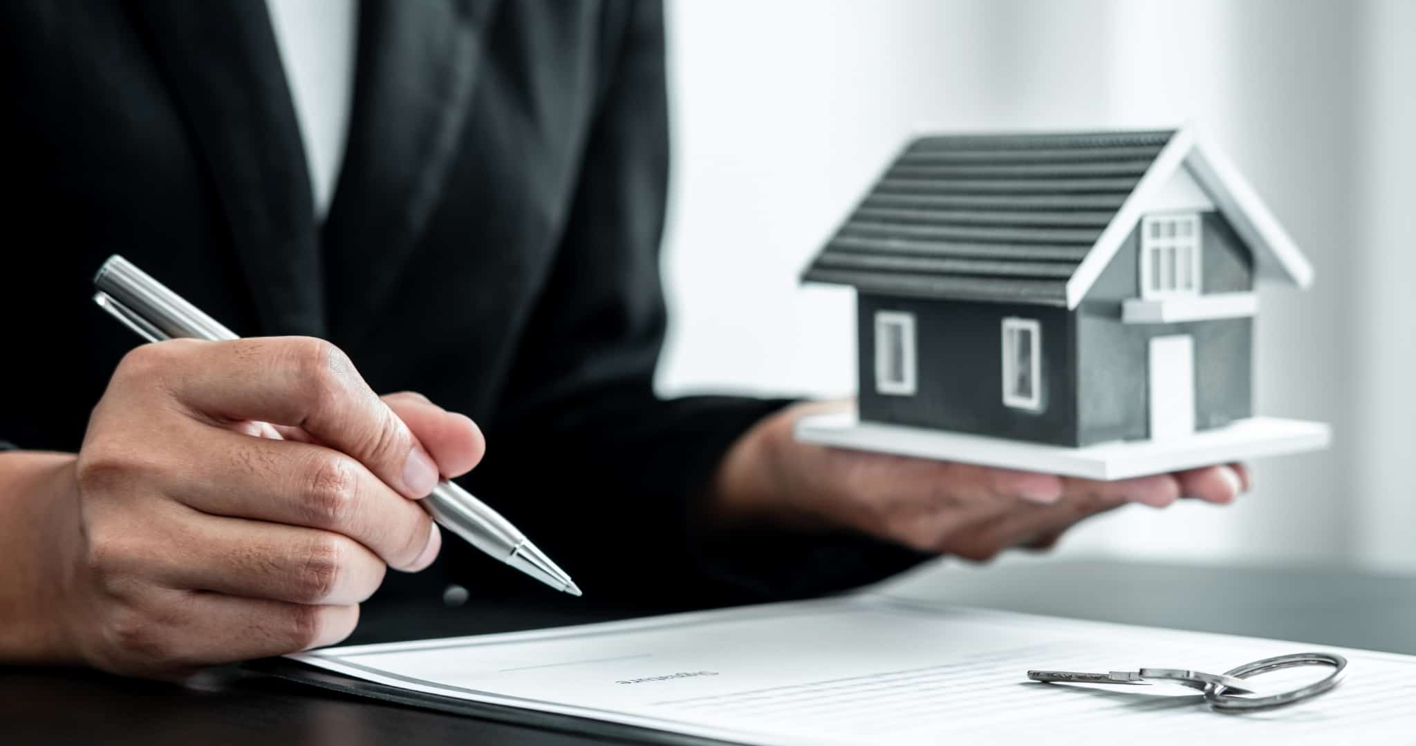 Homem de fato a assinar contrato revisão crédito habitação com caneta e casa miniatura nas mãos