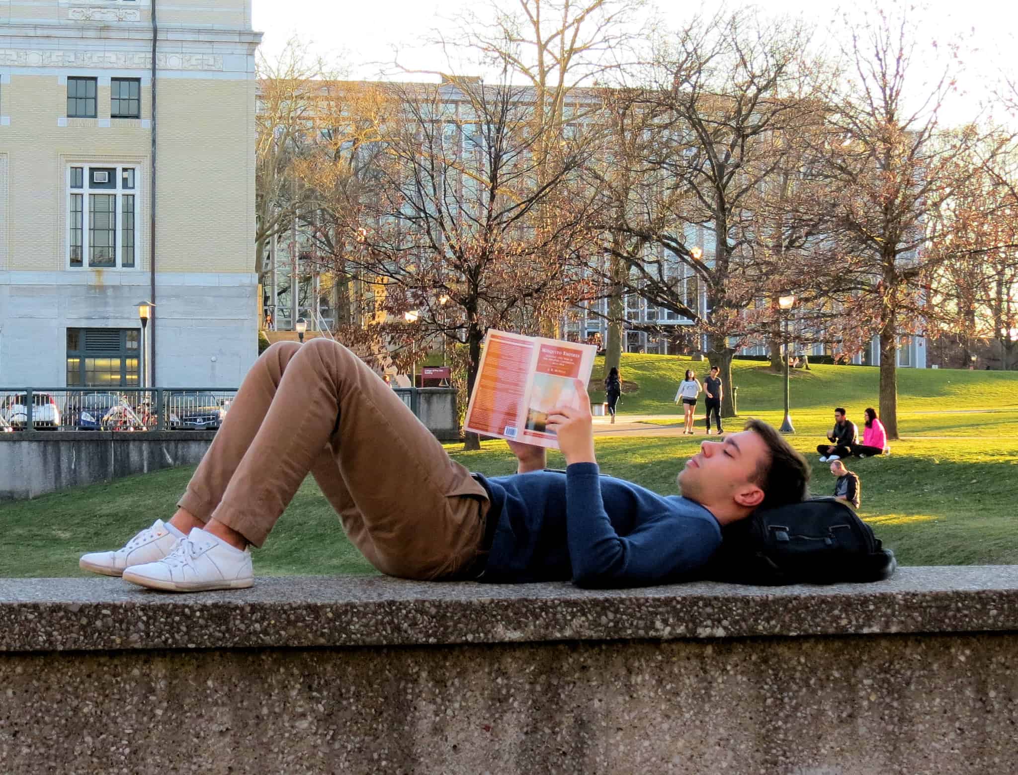 jovem estudante deitado em cima de um muro , junto à universidade, a ler um livro, com a cebça em cima da mochila