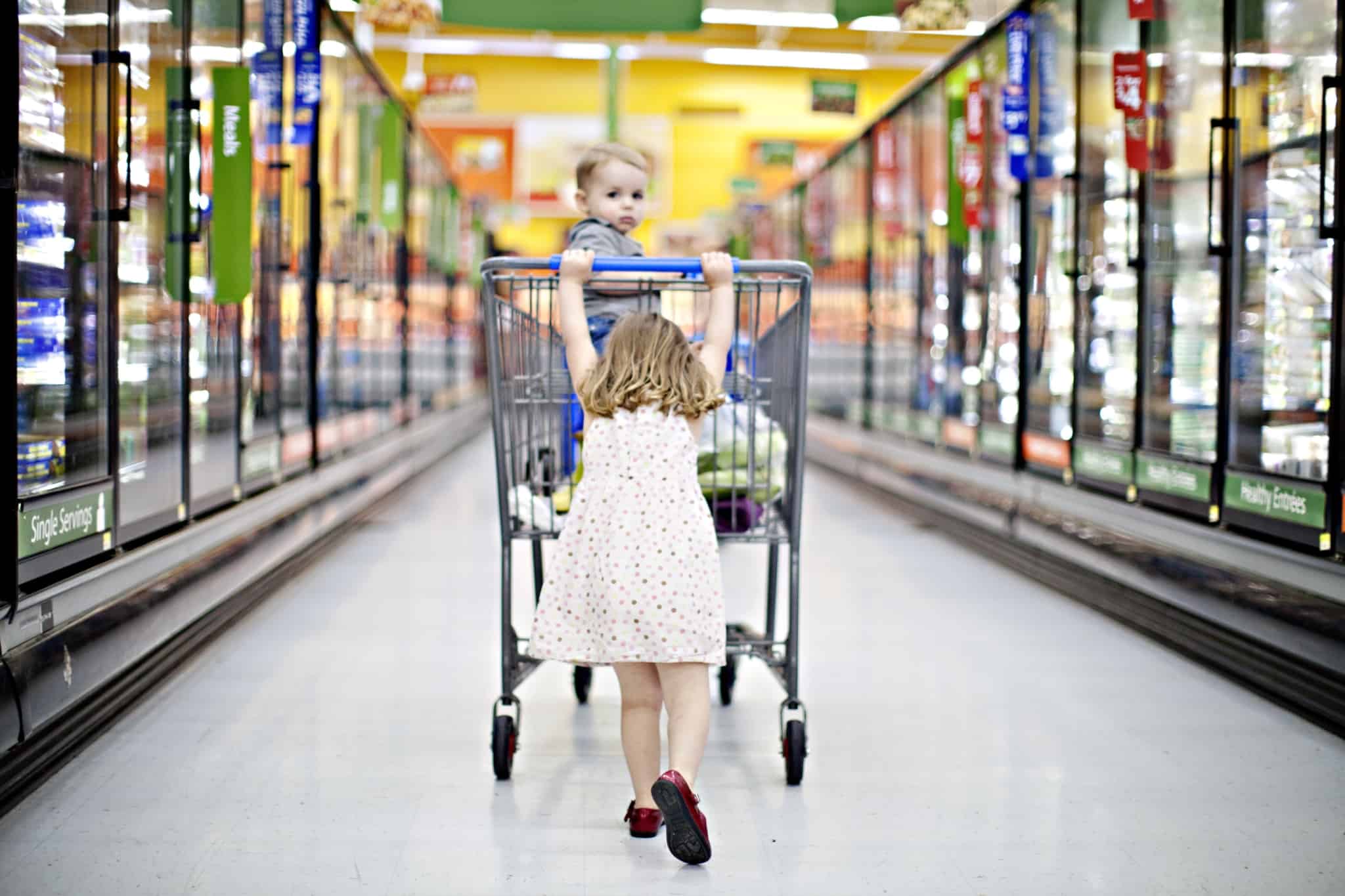 para ilustrar os hábitos de consumo do seu filho, a imagem mostra, num corredor de um supermercado, uma menina empurra um carrinho de compras com o pequeno irmão lá dentro