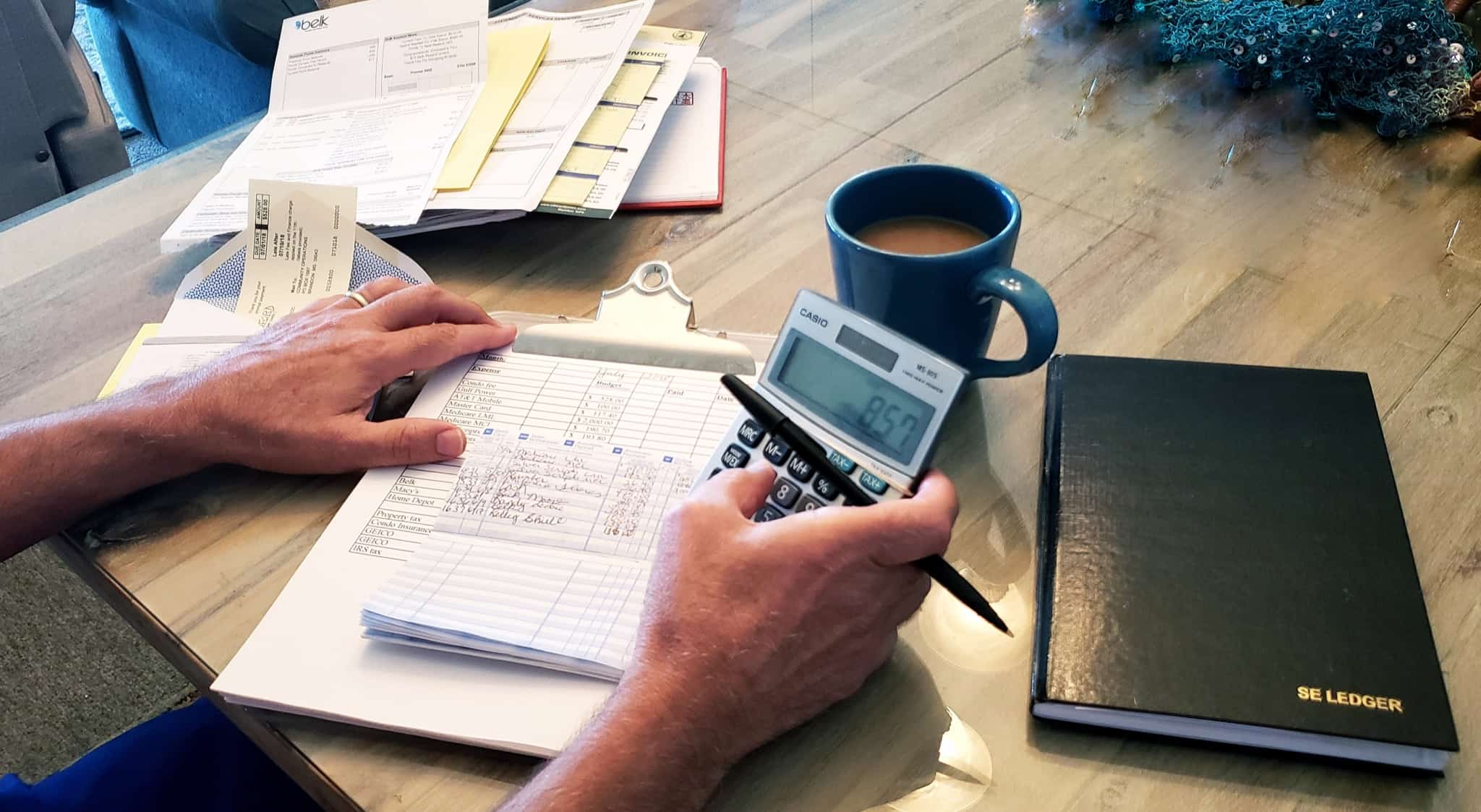 jovem homem faz calculos, recorrendo a uma calculadora, analisando um série de contas espalahdas em cima da sua mesa