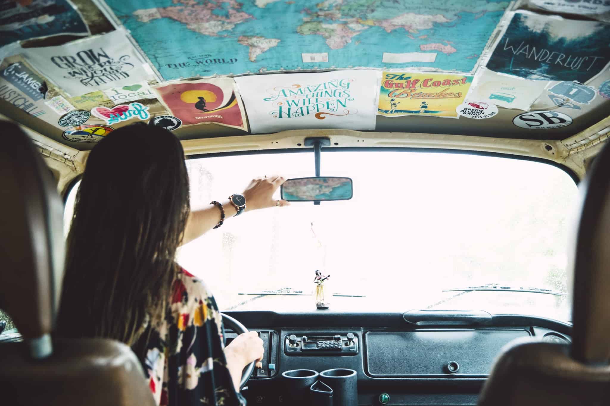 jovem mulher, ao volante de uma carrinha, ajeita o espelho retrovisor para partir na sua viagem de carro pela Europa.