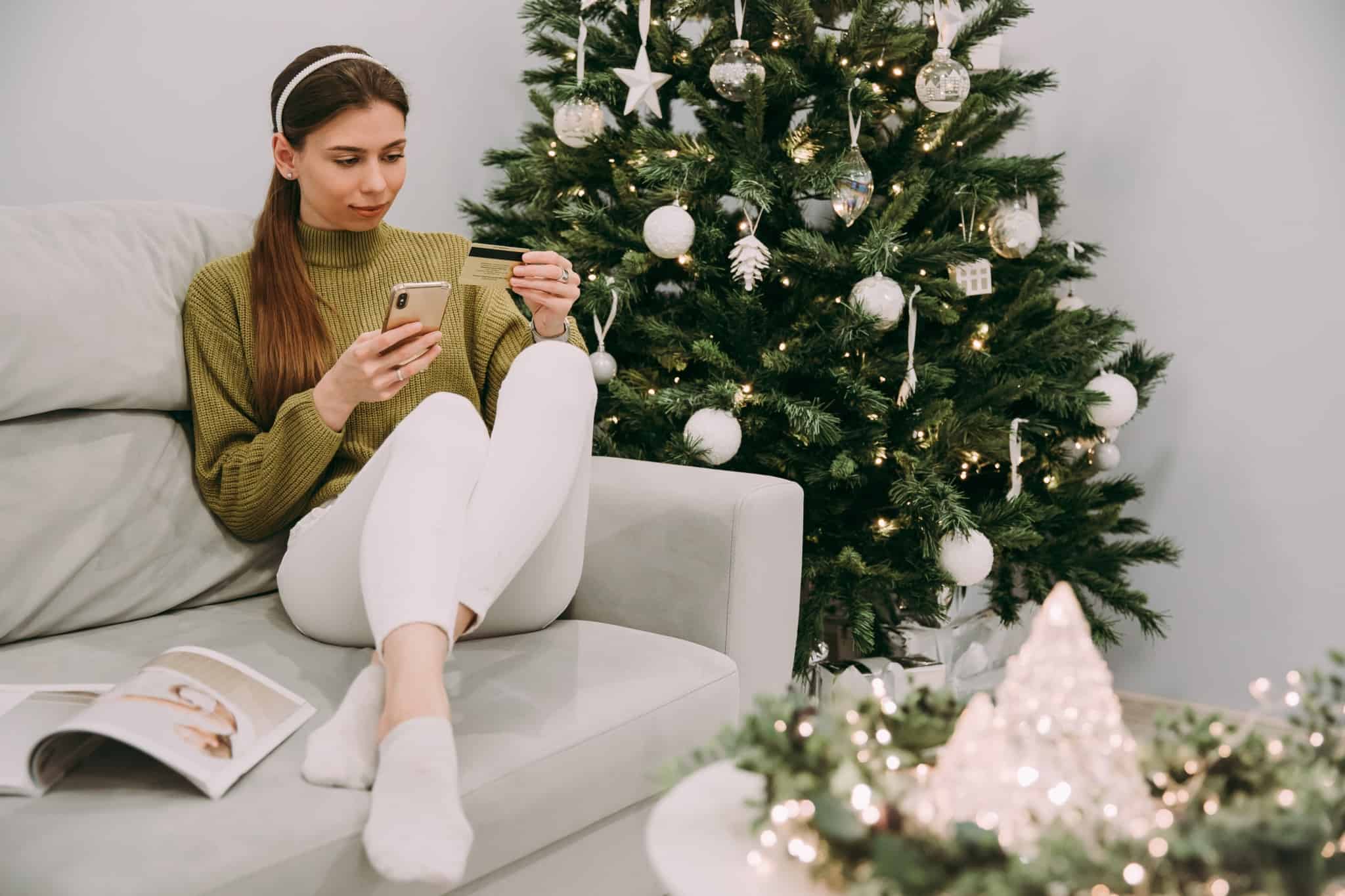 jovem mulher, junto à árvore de Natal, faz compras por telemóvel, com pagamento por cartão crédito