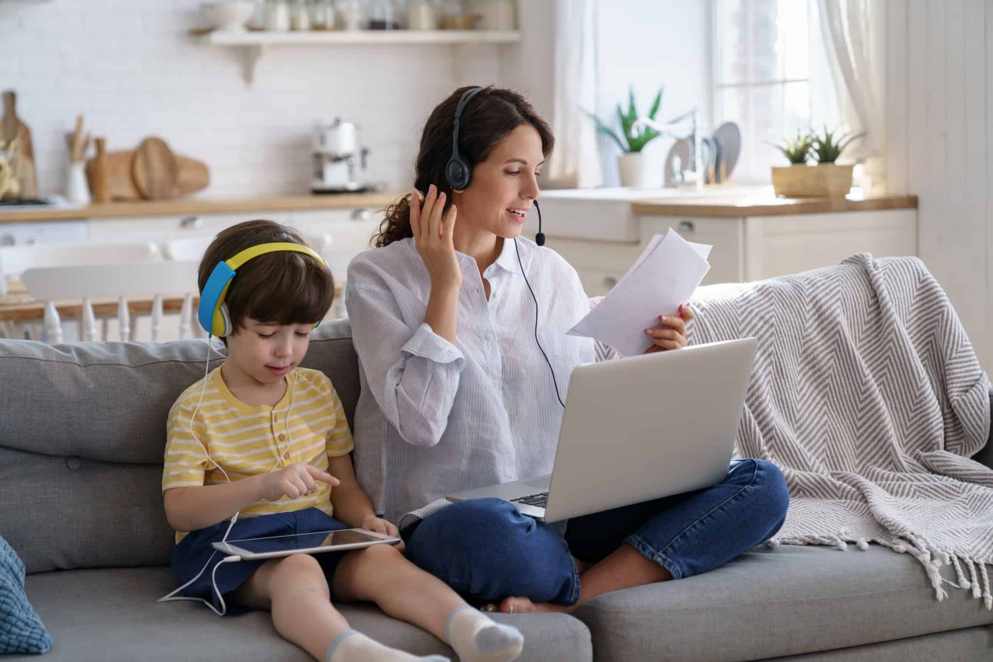 jovem mãe trabalha em teletrabalho, está no sofá, ao lado do seu pequenbo filho que está de phones e a dar o tablet enquanto a mãe fala ao telefone e consulta o computador