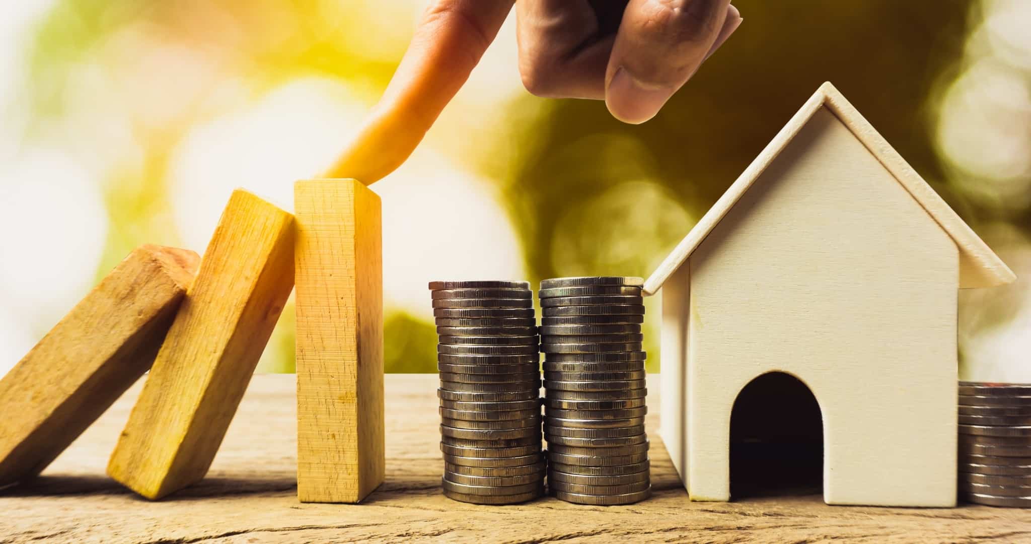 Três peças de domino a cair para cima de moedas e casa de madeira a simbolizar casa destruída protegida por seguro multirriscos