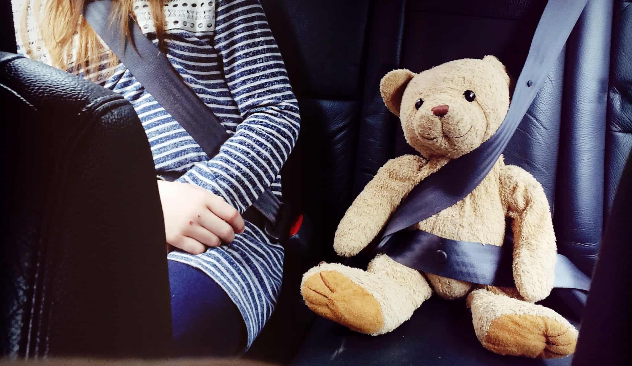 criança sentada no carro de cinto de segurança posto. Ao seu lado segue um urso de peluche de cinto posto