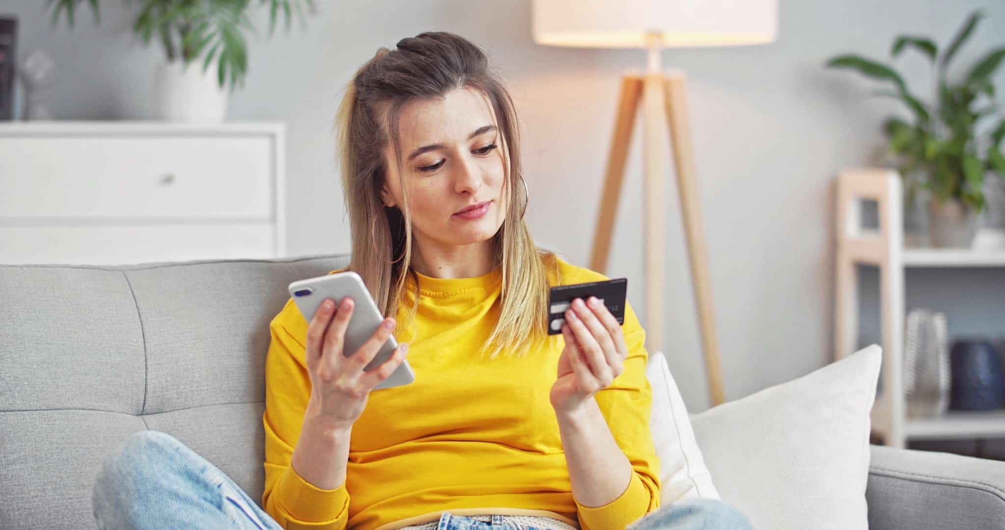 jovem mulher faz compras através do telemovel, sentada no sofá de casa, usando o cartão de crédito